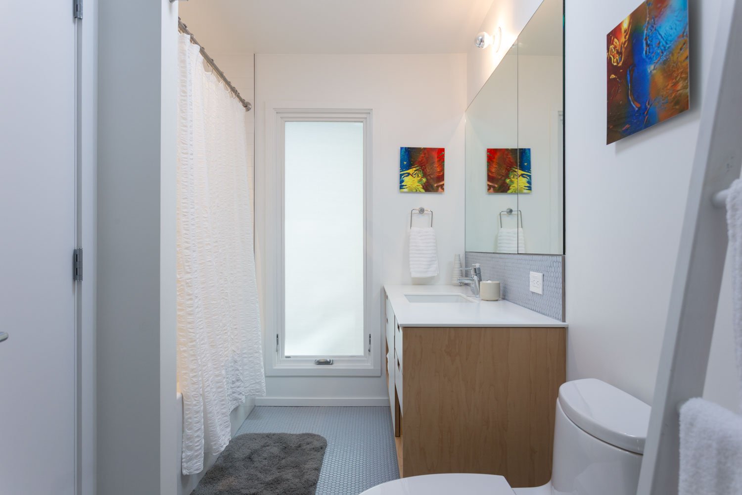 Modern bathroom with white walls, a frosted glass door, a shower with a white curtain, a wooden vanity with a white countertop, a rectangular sink, a large mirror, two small colorful abstract paintings, a towel hanging on a hook, a toilet, and a gray