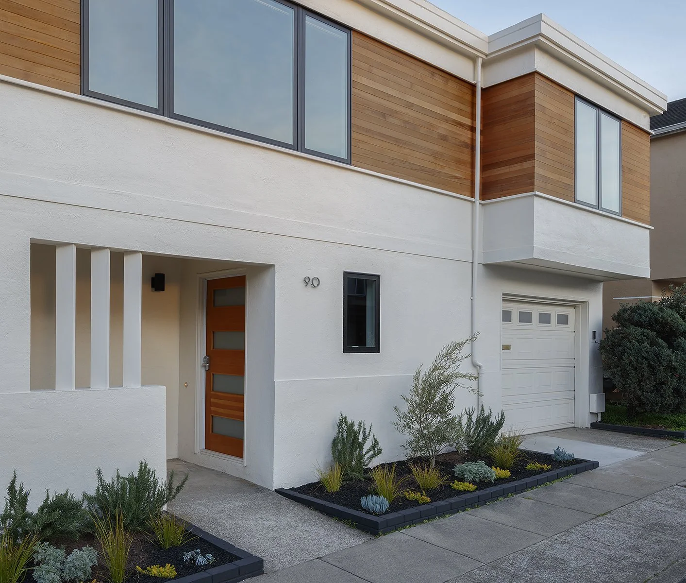 Modern two-story house with a white and wood exterior, featuring a front door, small garden bed, and large windows.