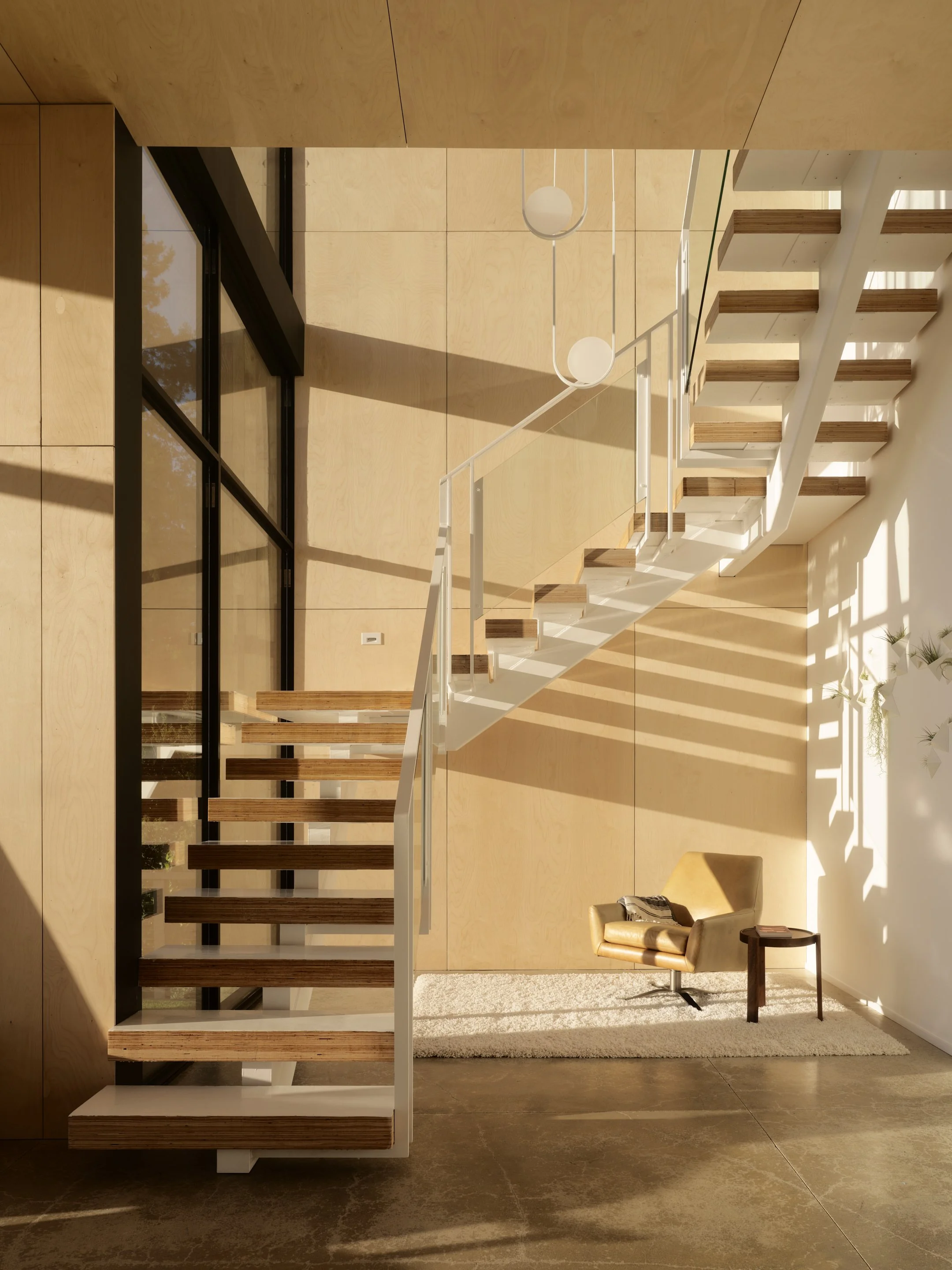 Modern interior space featuring a floating staircase with wood and black metal steps, a white railing, a beige armchair with a throw blanket next to a small wooden side table, a textured beige rug, and a large window casting shadows and sunlight into