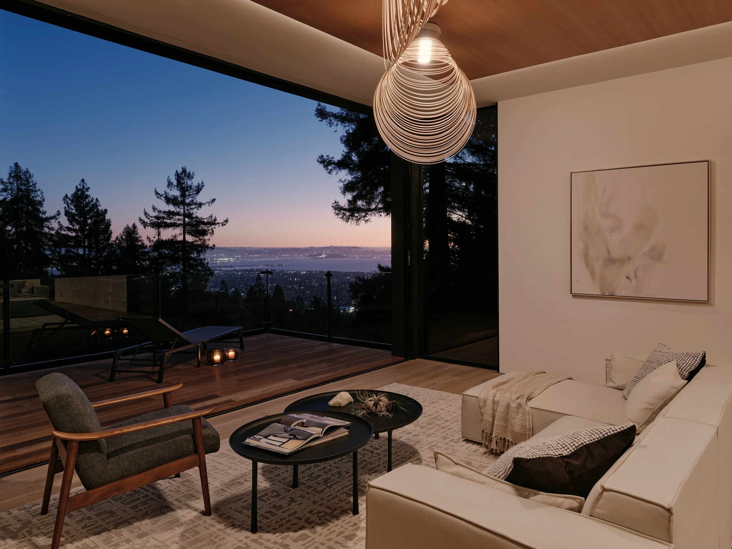 A modern living room with a cream sofa, patterned throw pillows, and a black coffee table with magazines. There are two armchairs, a rug, and a large window showing a scenic view of trees, a river, and a city skyline at dusk. A unique pendant light h