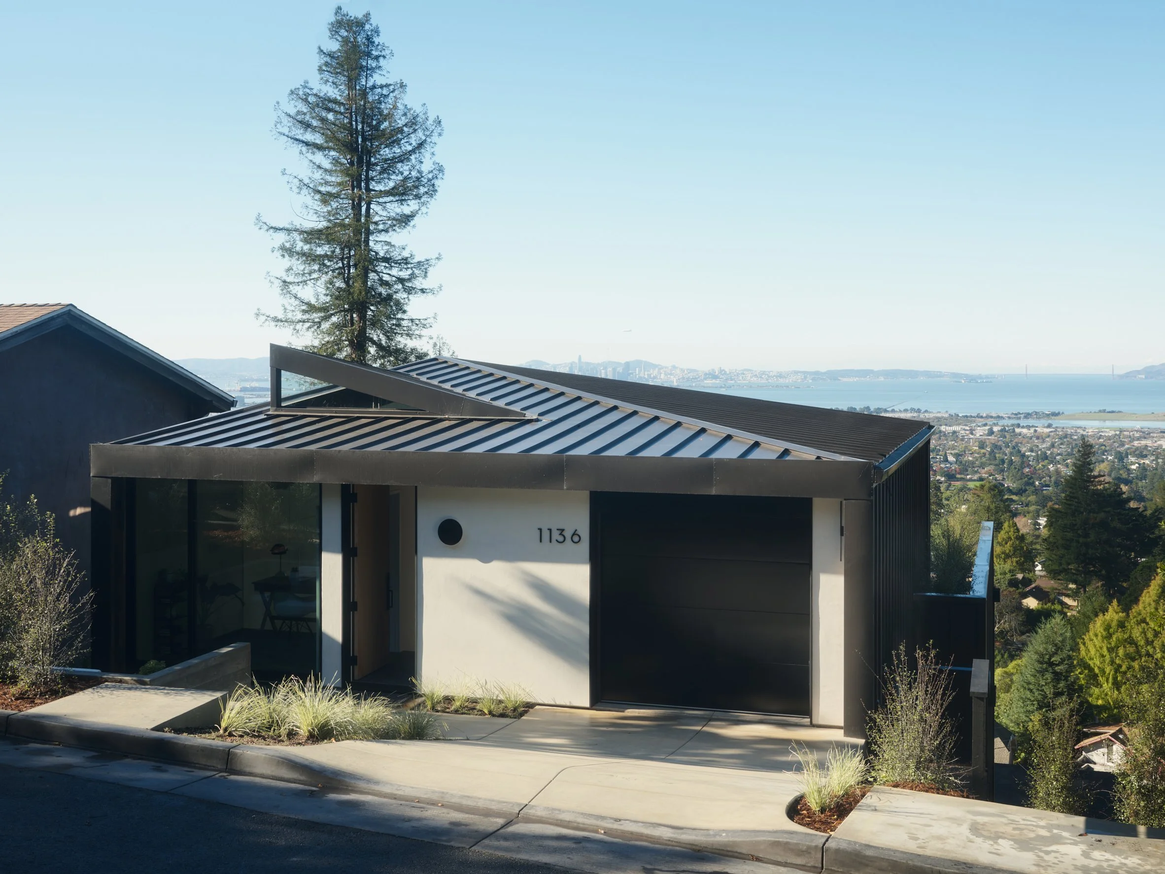 Modern house with black and white exterior, metal roof, street number 1136, in a hilly area with trees and a view of the city and water in the background.