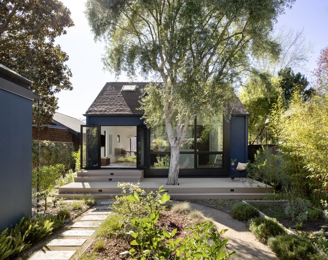 A modern house with large glass windows and a black exterior, surrounded by a lush garden with a pathway, trees, and shrubs, under a clear blue sky.