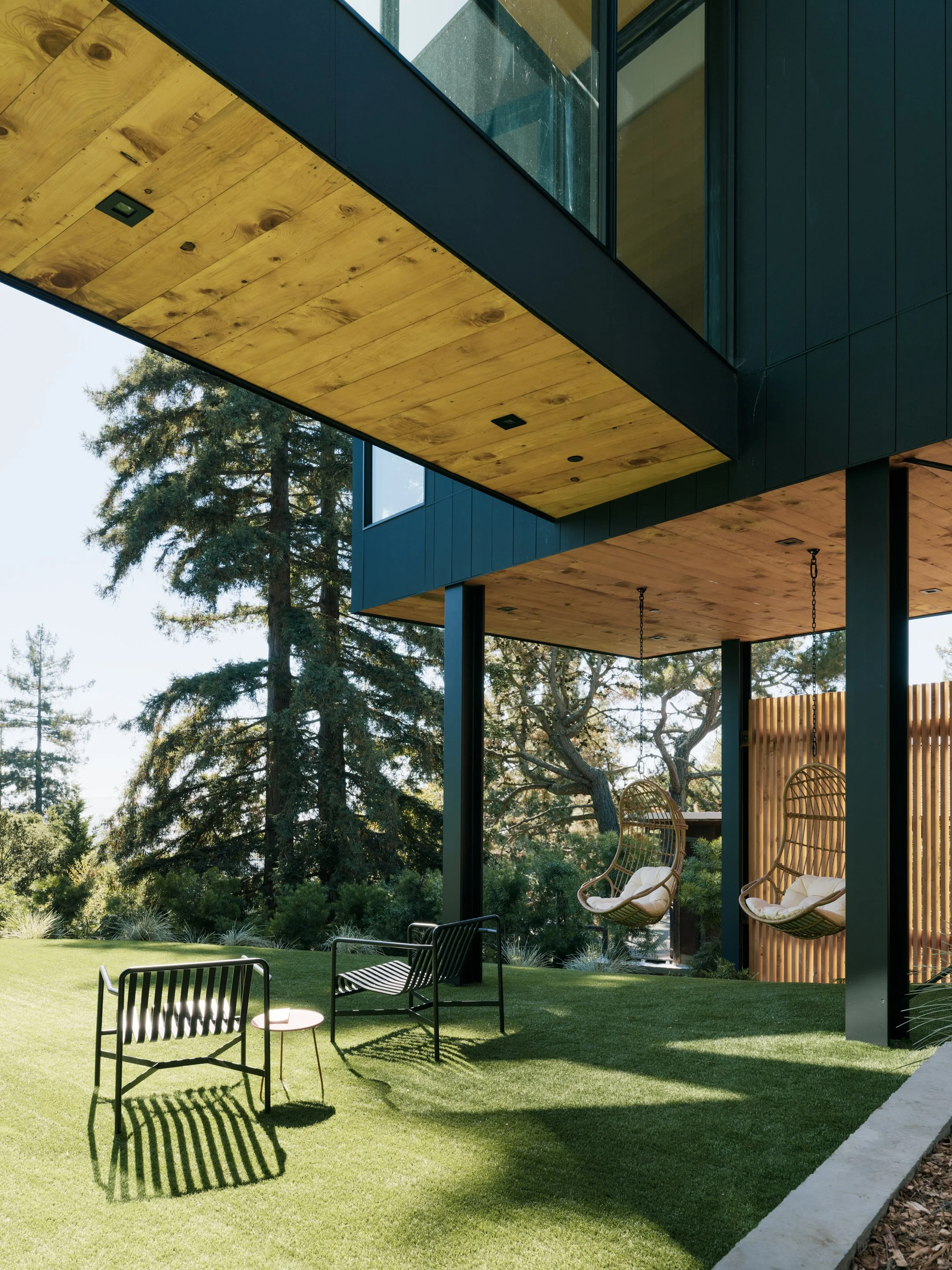 Modern house with an overhanging balcony featuring wooden paneling, large glass windows, and outdoor seating including hanging chairs, set in a lush green yard with trees in the background.