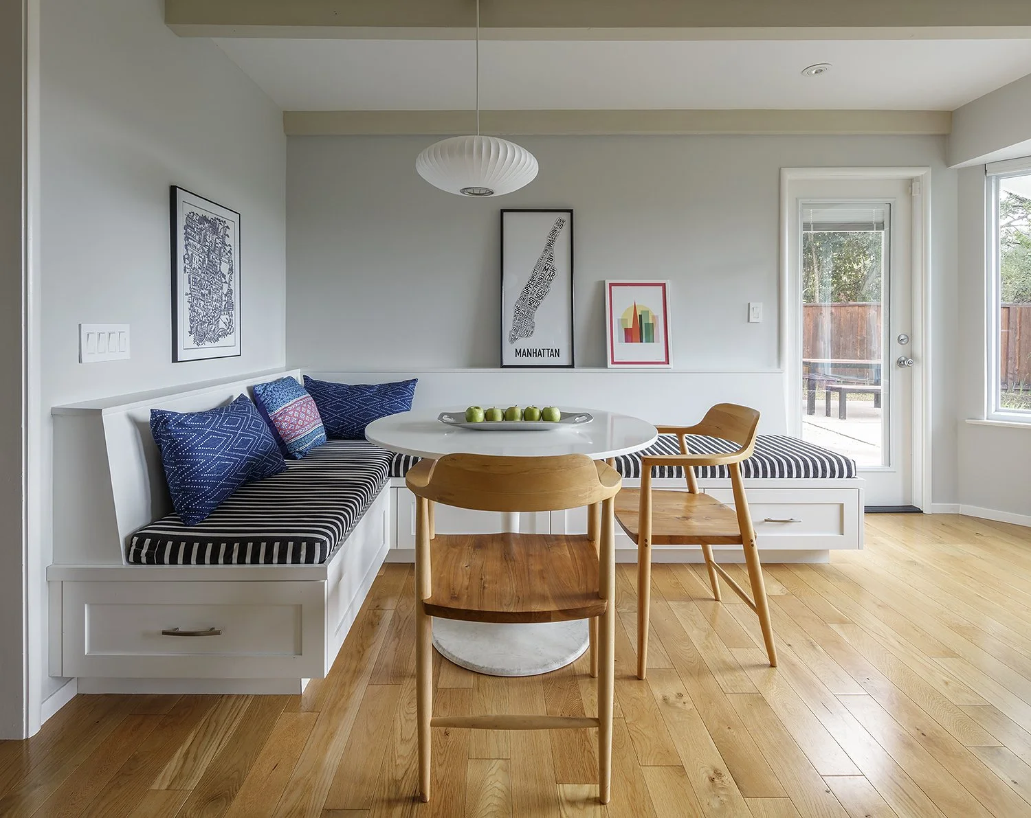 A cozy dining area with a built-in bench seat with striped cushions and pillows, a round white table with a tray of green apples, two wooden chairs, wall art including a Manhattan map and colorful skyline, a hanging white pendant light, and a glass d