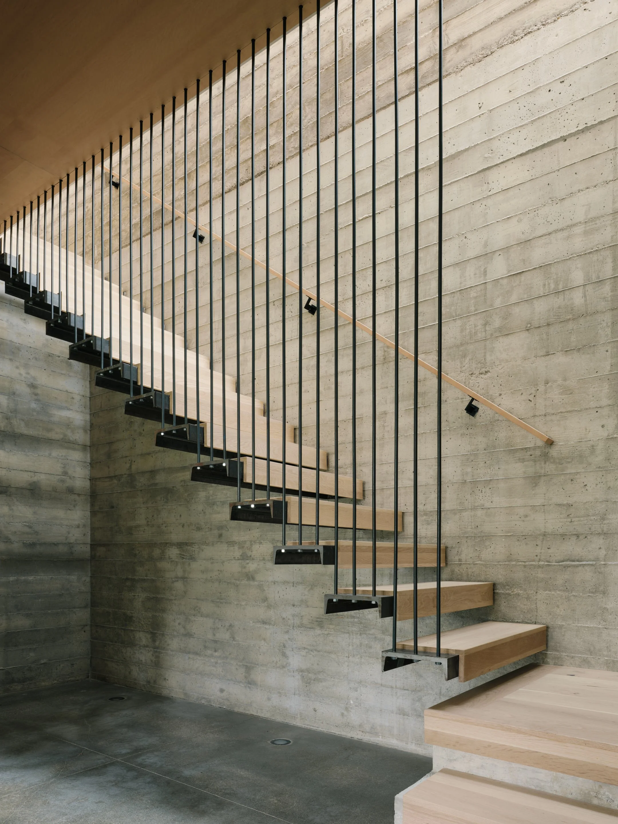 A modern staircase with black metal railings and light wooden steps against a concrete wall. The staircase is floating and minimalistic in design, with spotlights illuminating the wall.