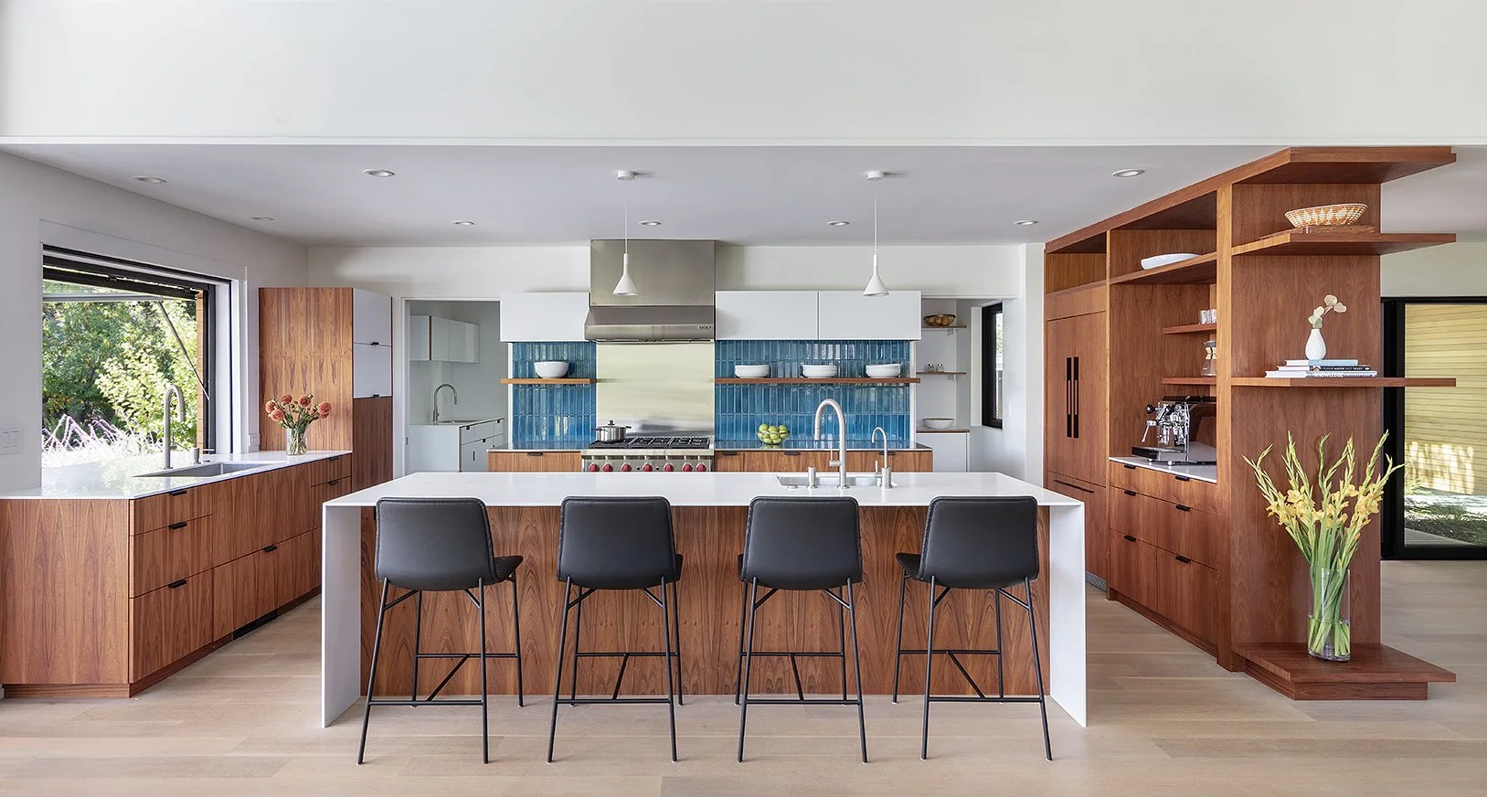Modern kitchen with wooden cabinetry, blue tiled backsplash, white island with black chairs, and open shelving with bowls and decor