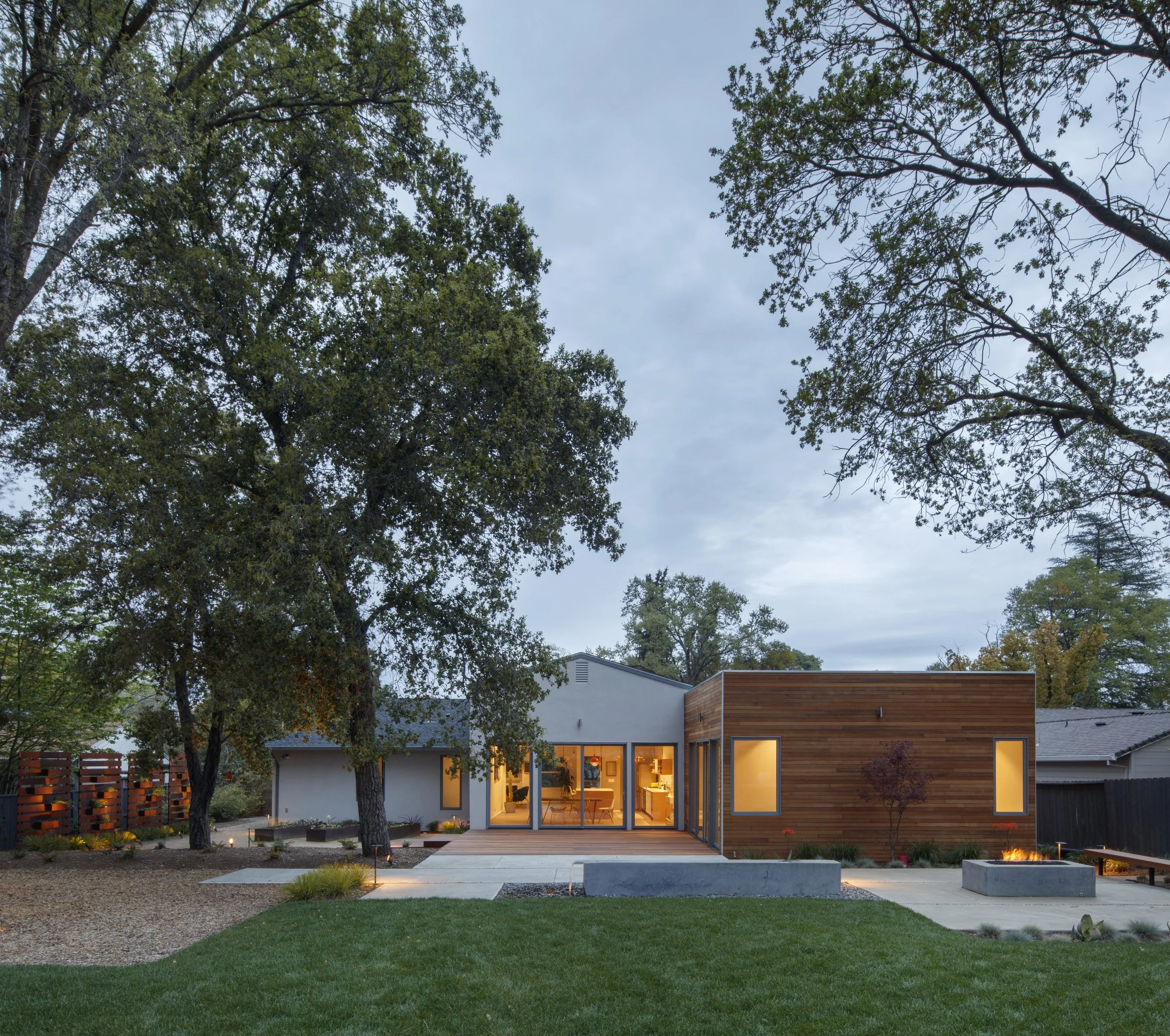 A modern house with large glass doors and wooden exterior panels, surrounded by trees and a well-maintained garden in the evening with the interior lights on.