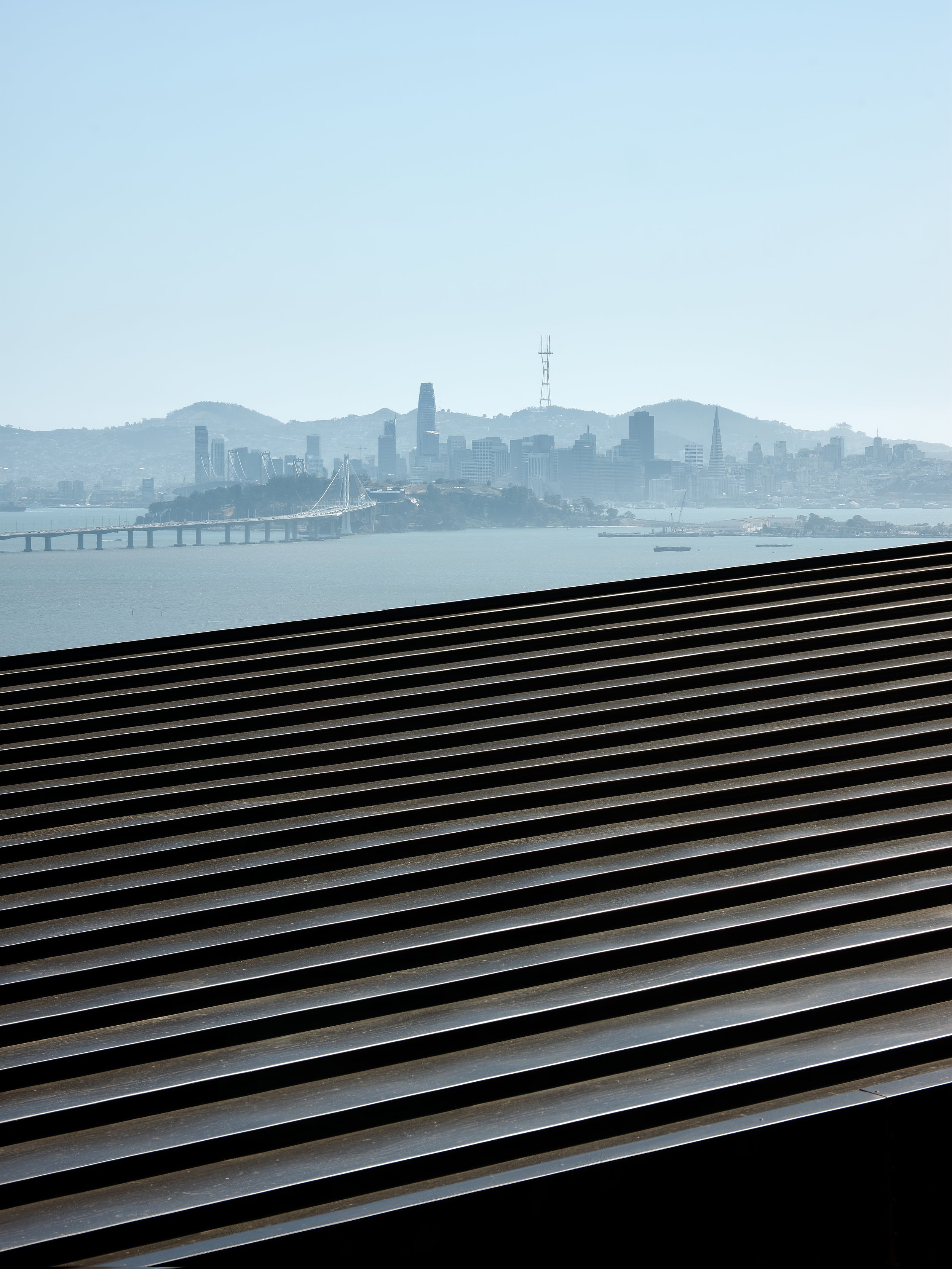 View of San Francisco skyline with the Bay Bridge in the foreground, viewed through slats, under clear sky.