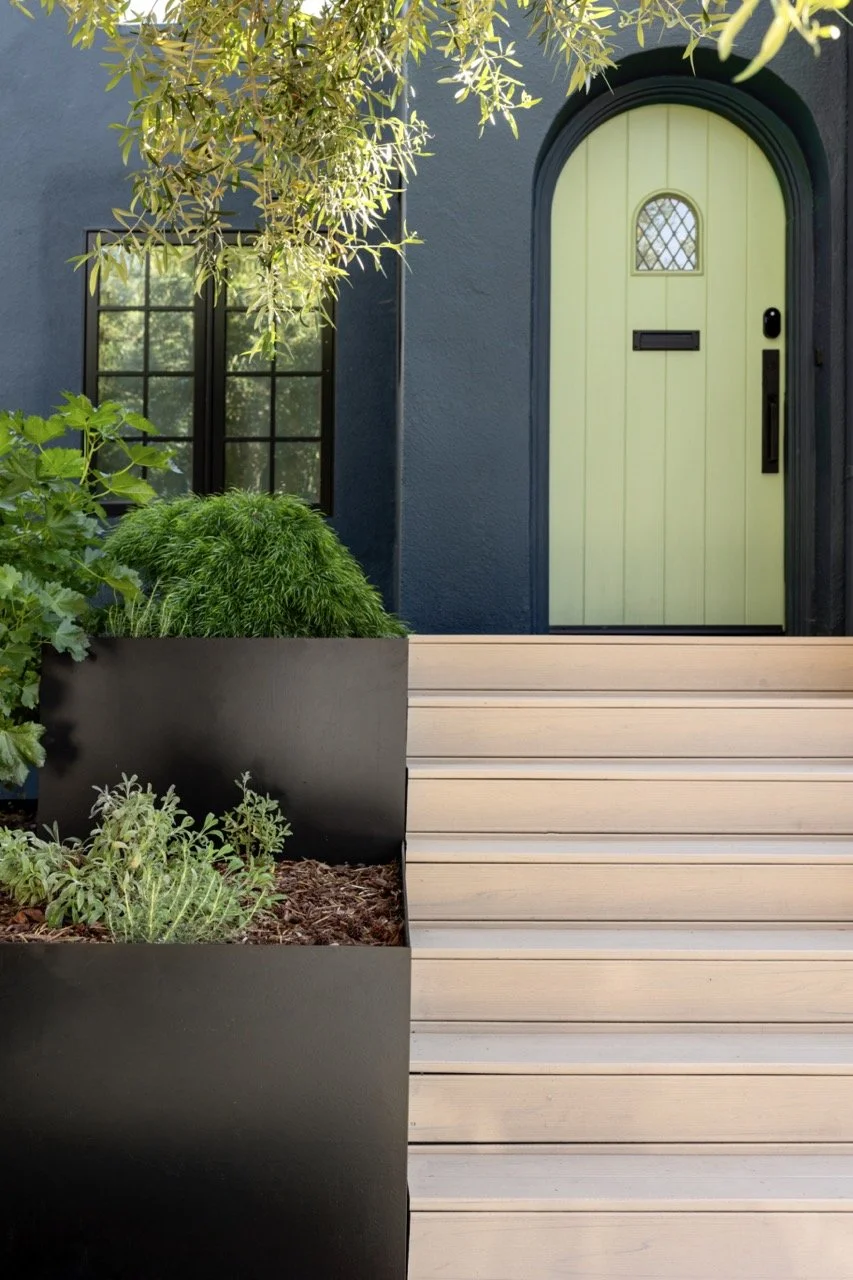 A front porch of a house with a lime green arched door, black hardware, a small window with a lattice, and a black mailbox. There are wooden steps, a black planter box with green shrubs, and a dark blue exterior wall with a window and overhanging tre