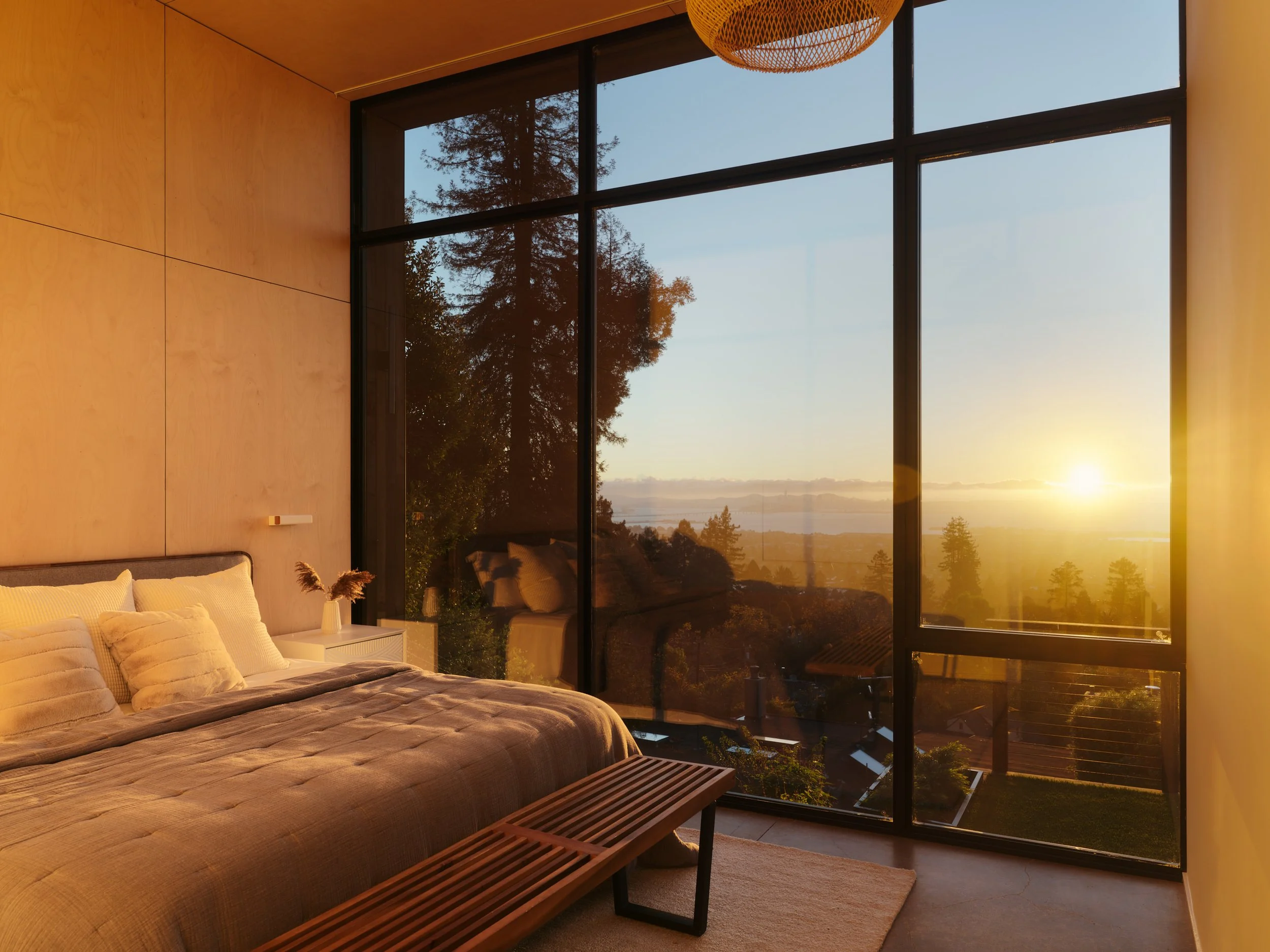 A bedroom with large floor-to-ceiling window showing a sunset view of trees and water. The bed has neutral-colored bedding and pillows, and there is a small bench at the foot of the bed.