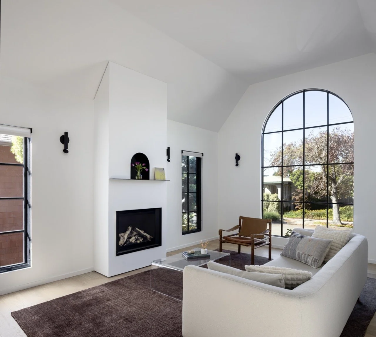 Living room with large arched window, white walls, modern fireplace, beige sofa with pillows, wooden chair, glass coffee table, and brown rug.
