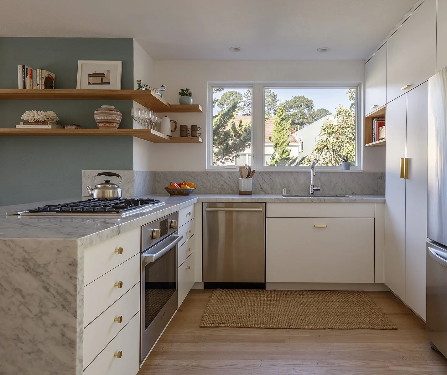 Modern kitchen with white cabinets, marble countertops, and open shelves, large window with a view of trees outside, and stainless steel appliances.