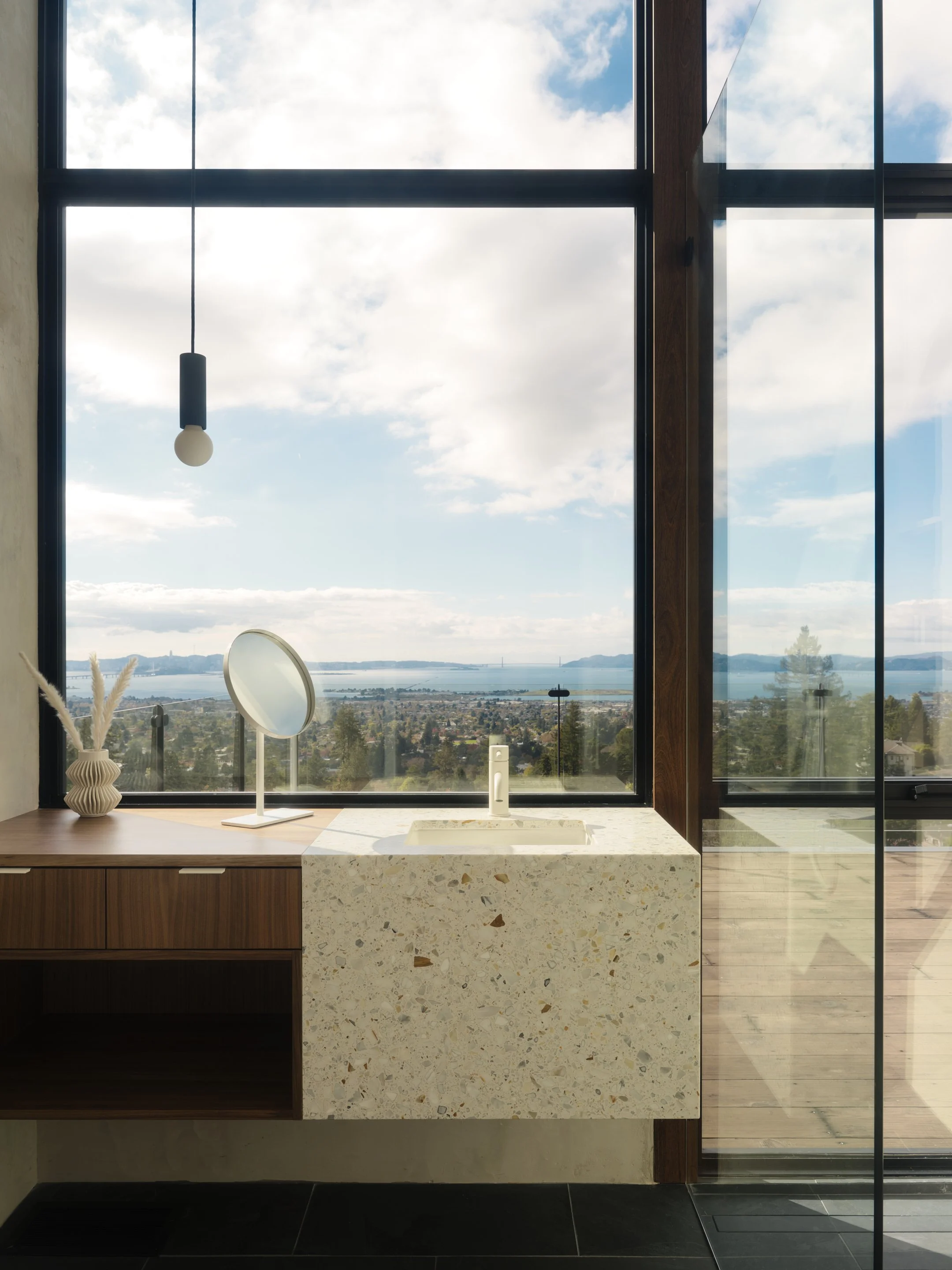 Interior view of a modern space with large floor-to-ceiling windows showing a cloudy sky and distant landscape, a countertop with a white sink, a decorative vase with pampas grass, and minimalistic decor.