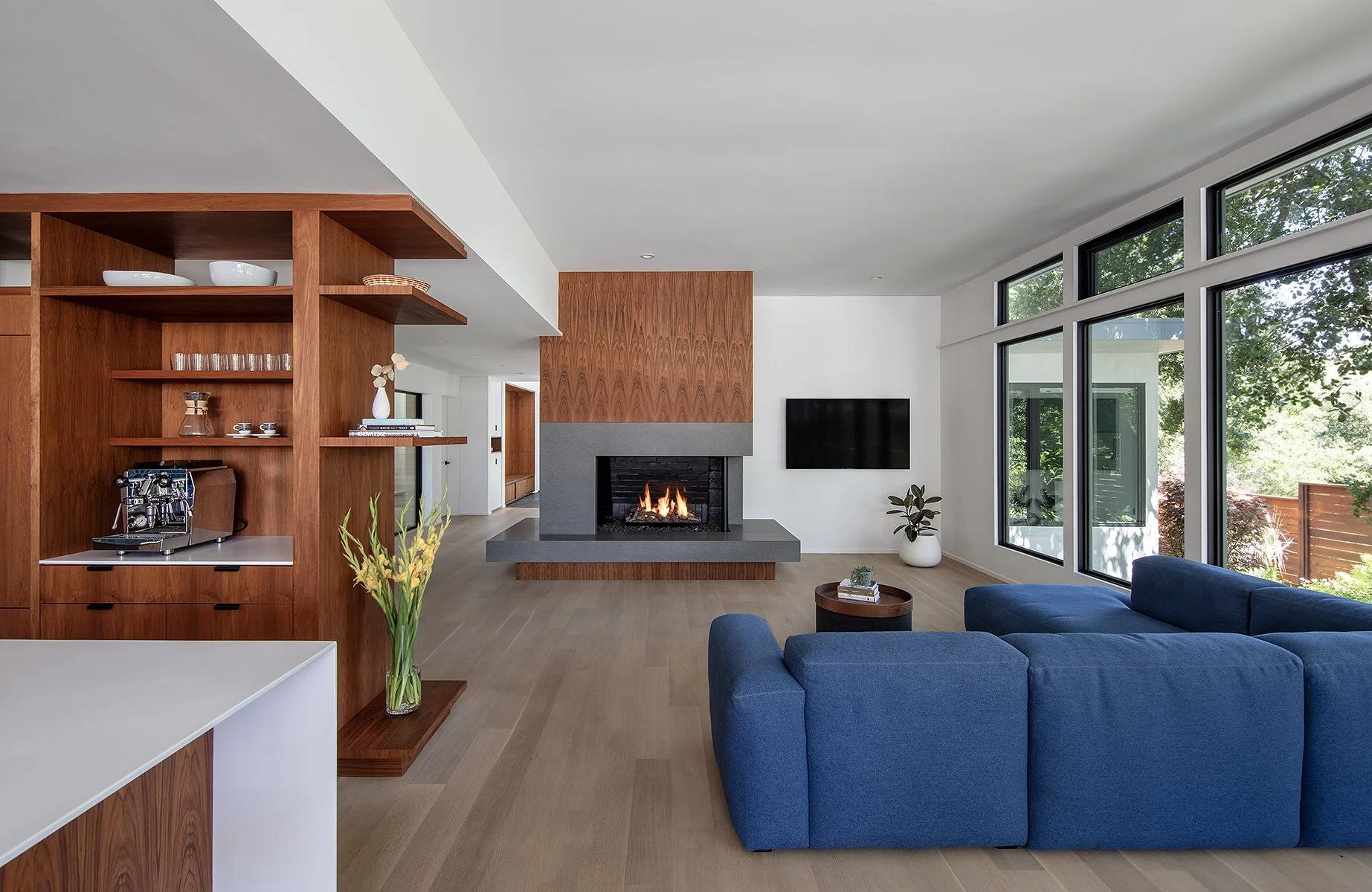 Modern living room with a fireplace, large windows, a blue sectional sofa, and wooden shelving.