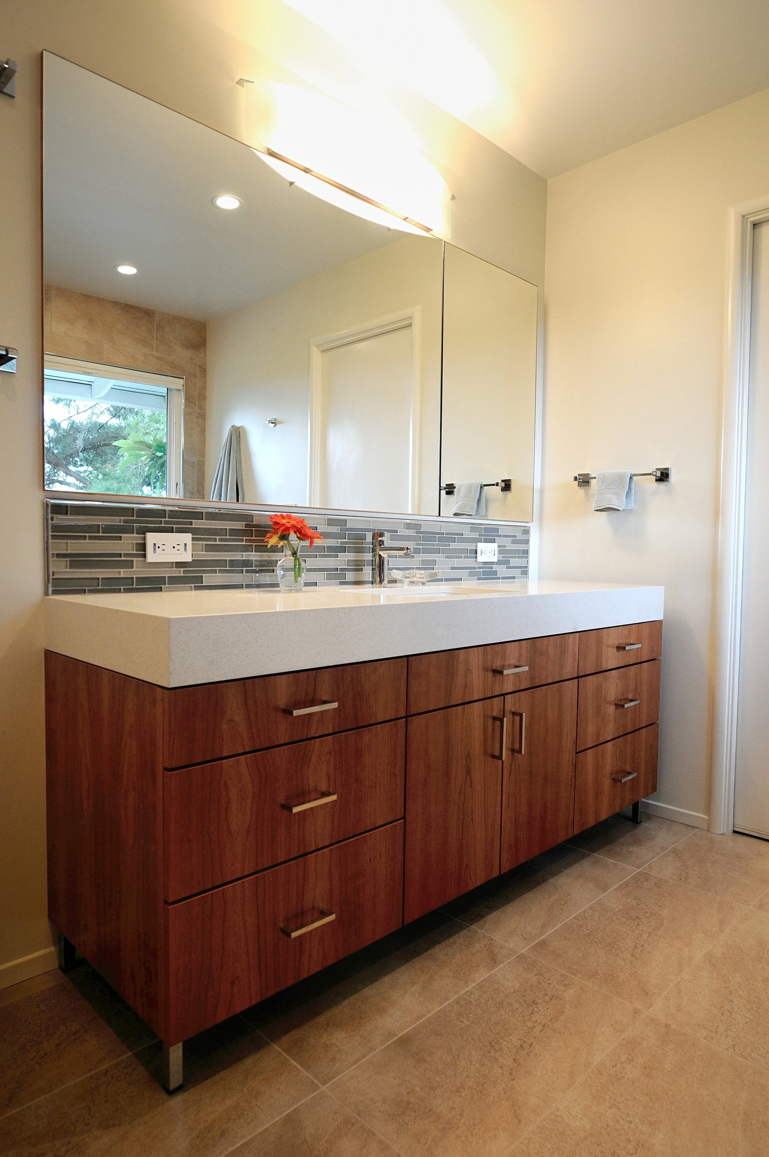 A modern bathroom vanity with a wooden cabinet, a white countertop, and a mirror above it. There is a small vase with a red flower, a faucet, and two electrical outlets. The wall has a decorative tile backsplash, and there are two towel racks with to