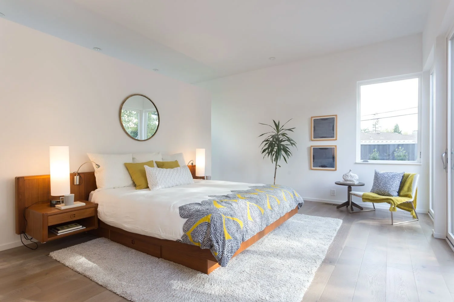 A modern bedroom with a wooden bed frame, white bedding, yellow and white pillows, two bedside lamps, a round mirror above the bed, a potted plant, a chair with a yellow blanket near the window, and two framed pictures on the wall.