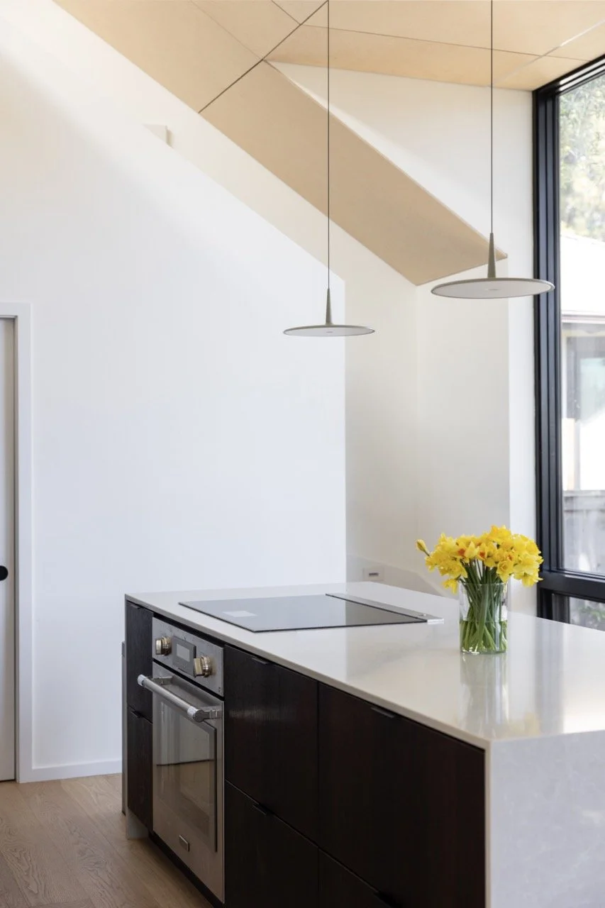 Modern kitchen with black cabinetry, white countertop, electric stove, glass vase with yellow flowers, large window, and minimalist pendant lights.
