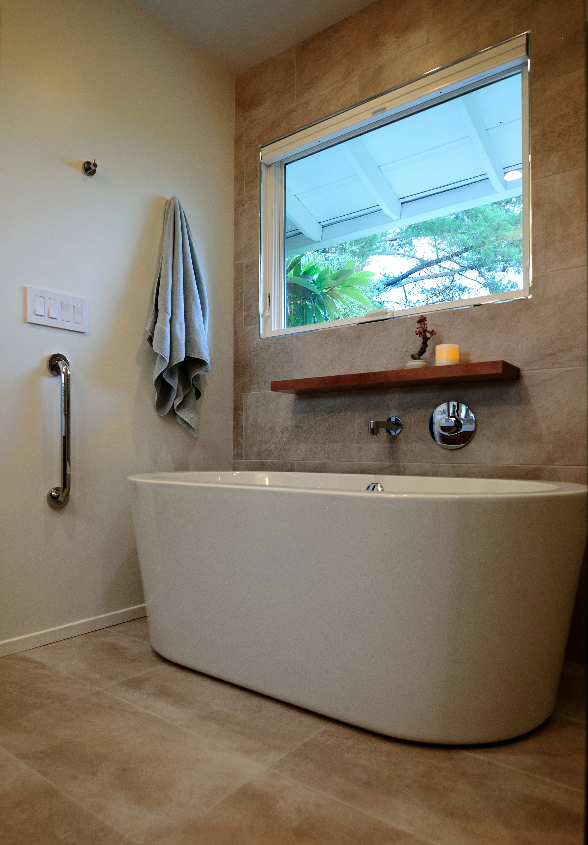 Modern bathroom with a white freestanding bathtub, a wall-mounted shower with a handheld showerhead, a towel hanging on a hook, and a window with a view of trees outside. The walls are tiled with beige tiles and the floor has matching tiles.