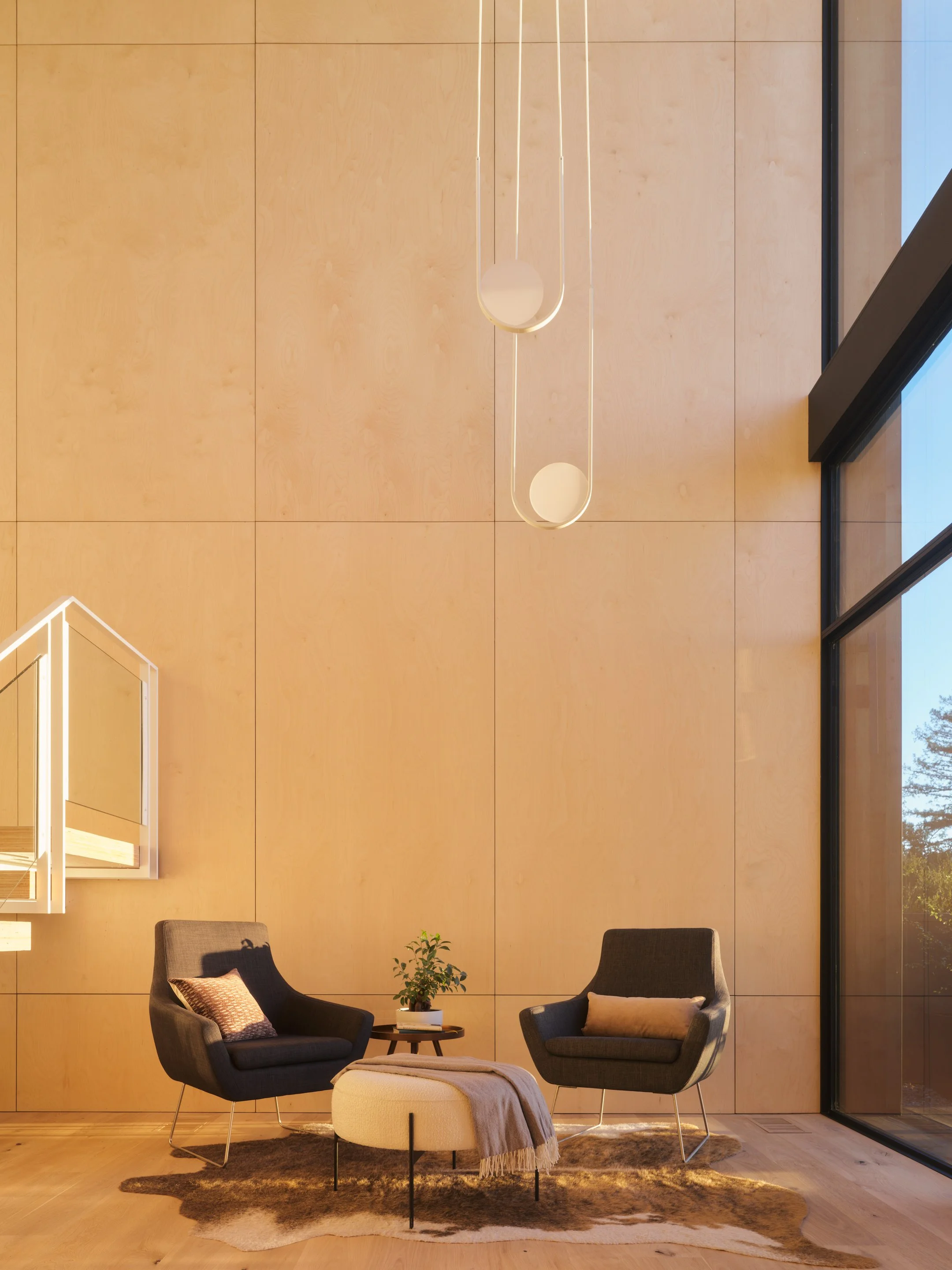 A modern living room with a high wooden wall, a round hanging light, two dark armchairs with pillows, a small coffee table with a plant, and a window showing trees outside.