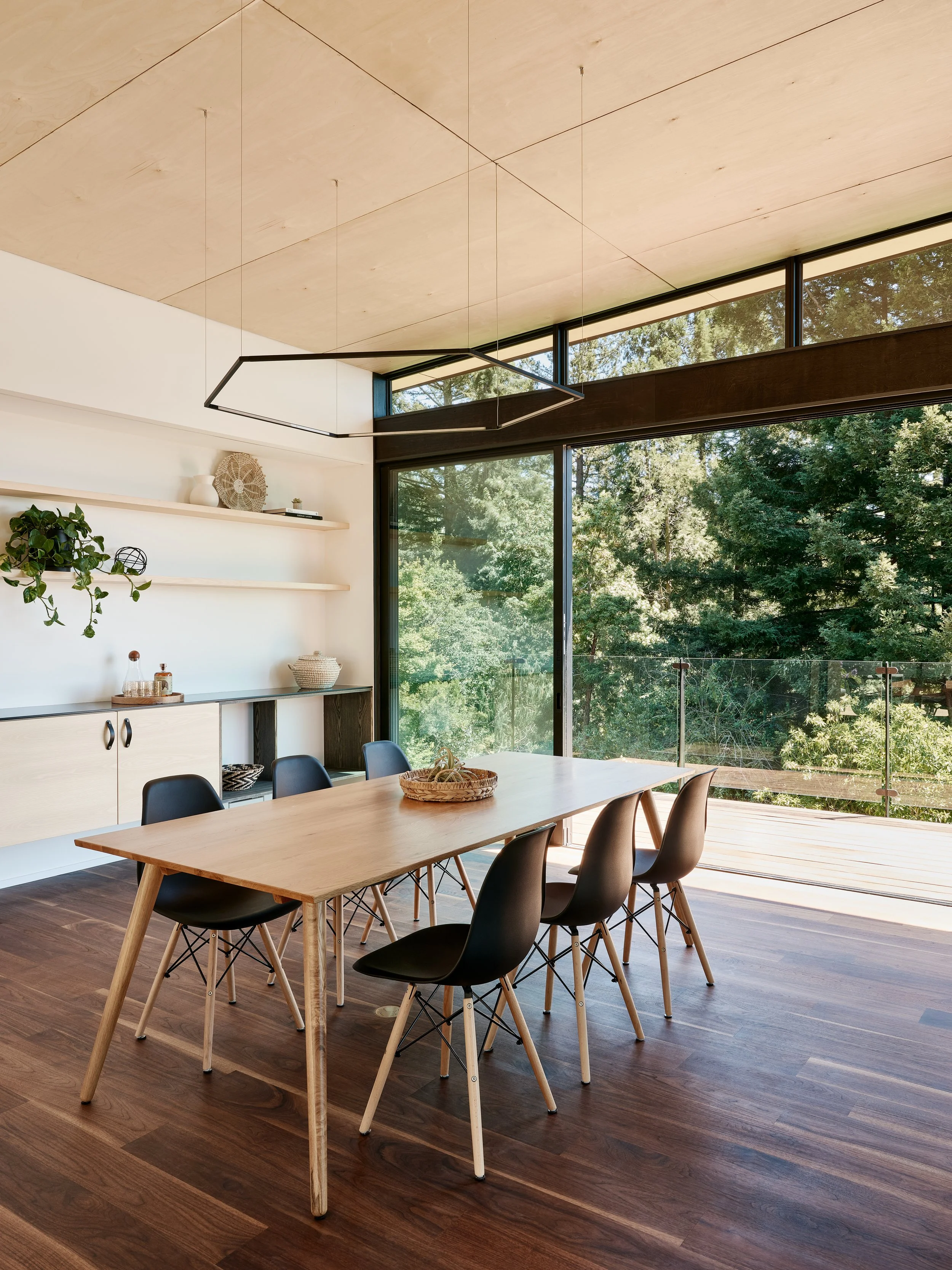Modern dining room with wooden table, black chairs, large windows showing trees outside, and a high ceiling with a minimalist black geometric chandelier.