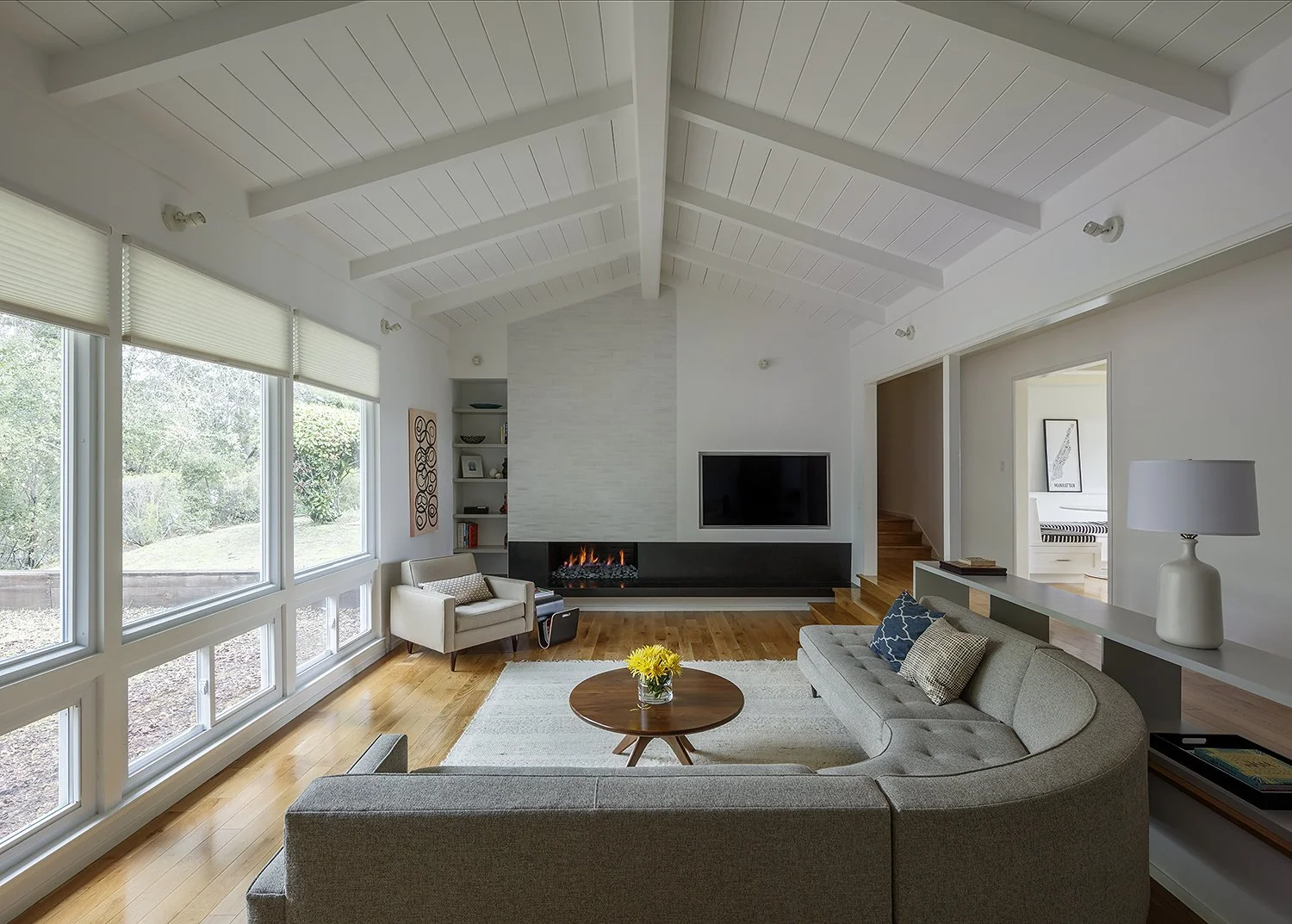 Living room with large windows, a white ceiling with wood beams, a fireplace, a flat-screen TV, and a gray sectional sofa with pillows.