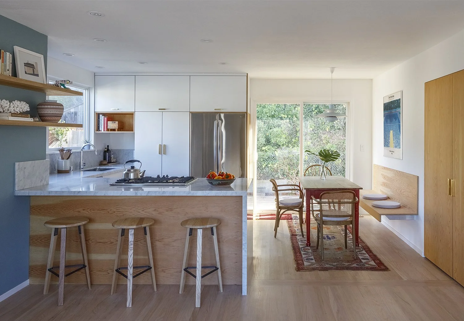 Modern kitchen with white cabinets, marble countertops, open wooden shelves, and large window showing trees outside.