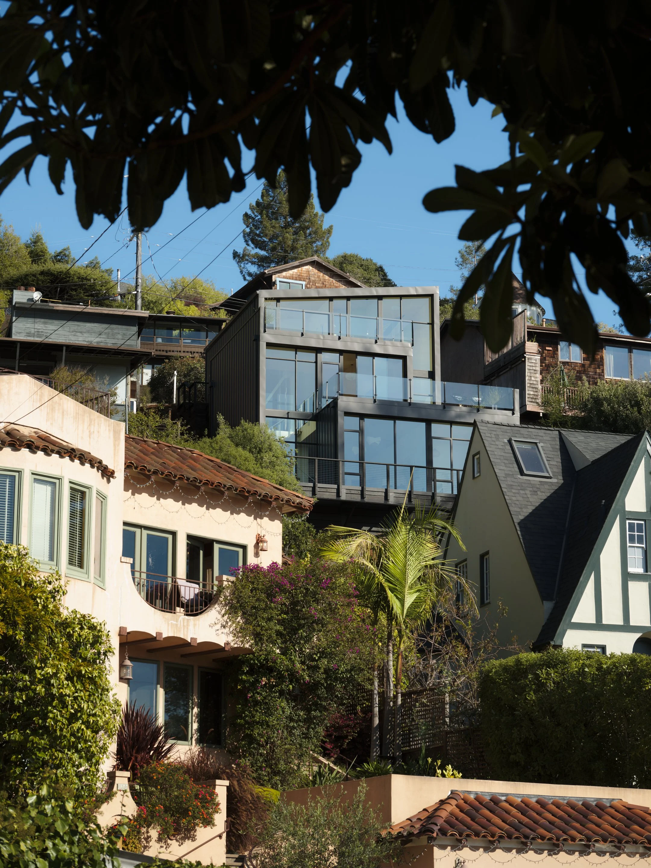 Multi-story houses with various architectural styles are built on a hillside, surrounded by greenery and trees, with a clear blue sky in the background.