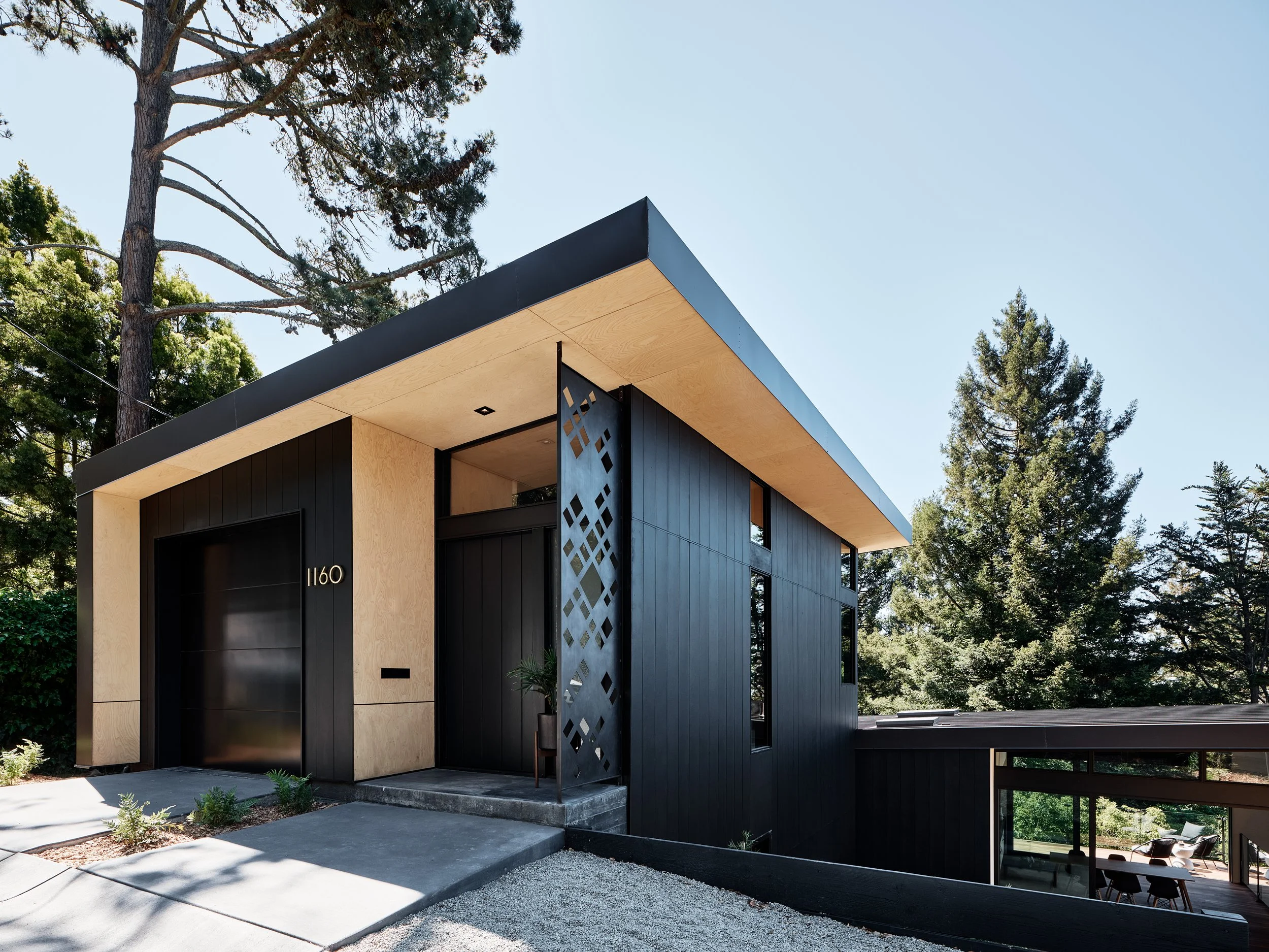 Modern two-story house with black and beige exterior, large black garage door, decorative metal screen, surrounded by tall trees and greenery, under a clear blue sky.