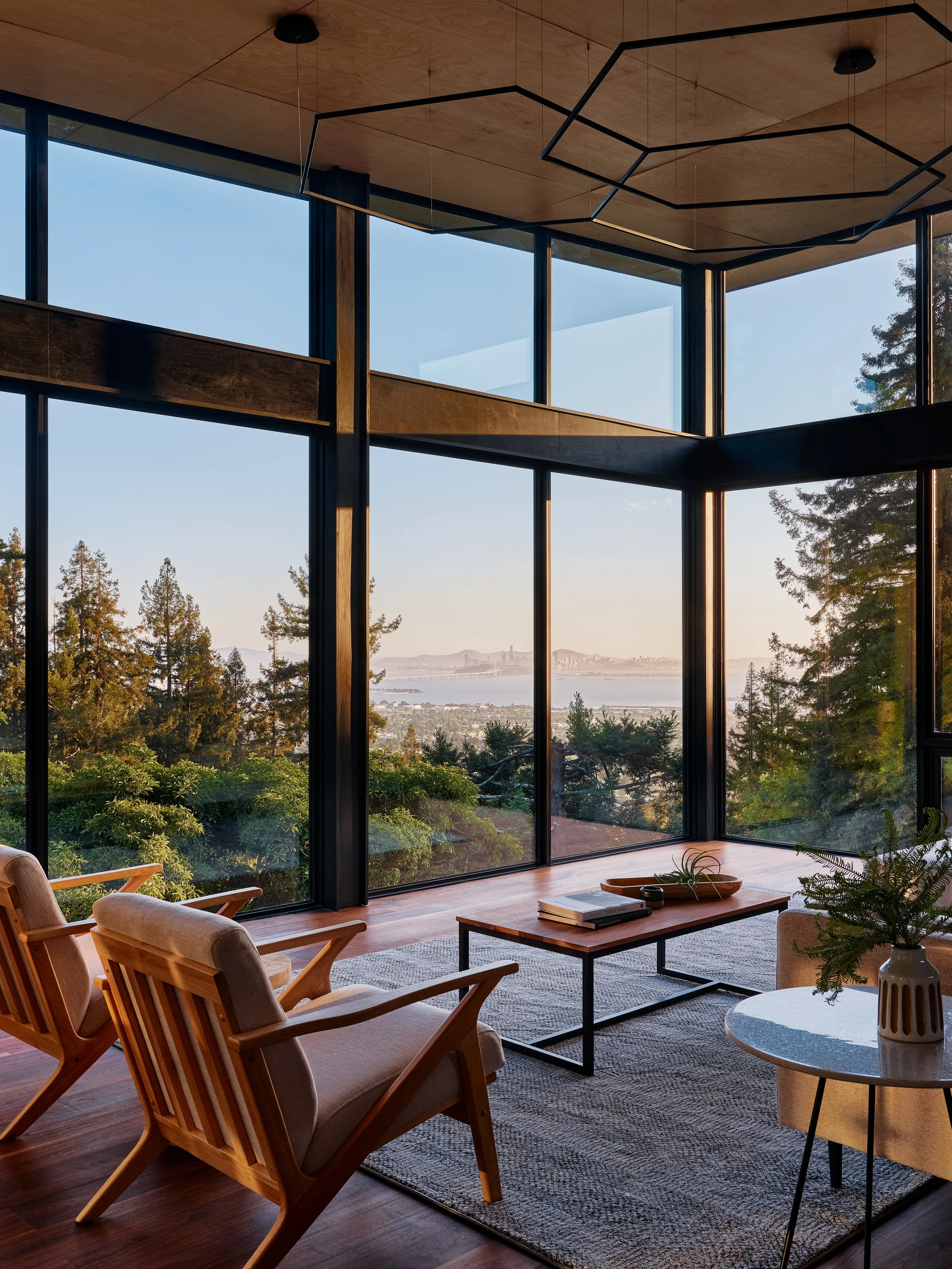 Modern living room with large floor-to-ceiling windows showcasing a scenic view of trees, water, and distant cityscape. The room features a coffee table, magazines, and cozy chairs with wooden frames.