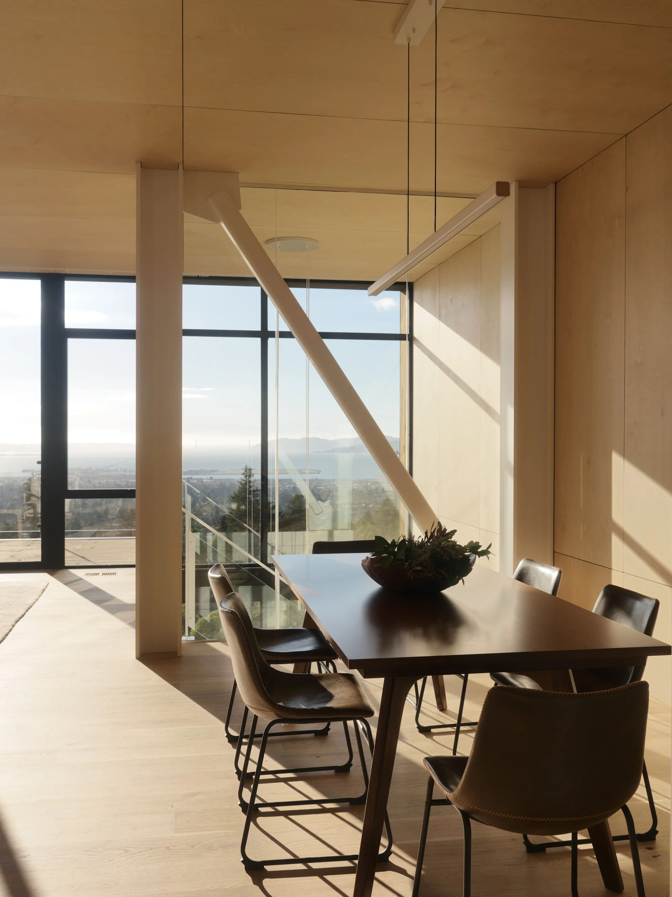 Modern dining room with wooden table and chairs, large windows offering a view of the landscape outside, and a decorative bowl with plants on the table. Bright sunlight and clear sky visible through the windows.