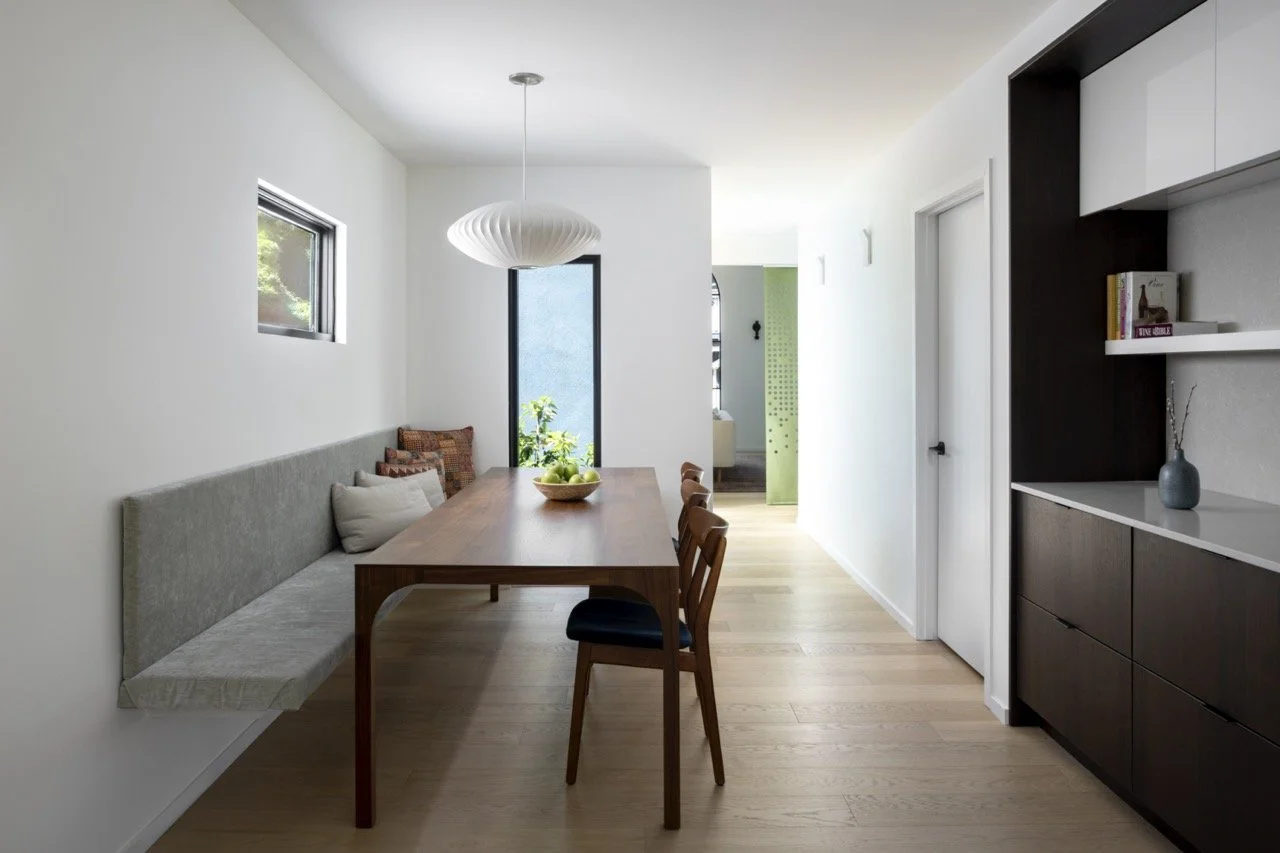 Modern dining room with a wooden table, a built-in bench with cushions, and two chairs, illuminated by pendant lighting, with a small window and a larger window behind the table, and a sideboard with decorative items.