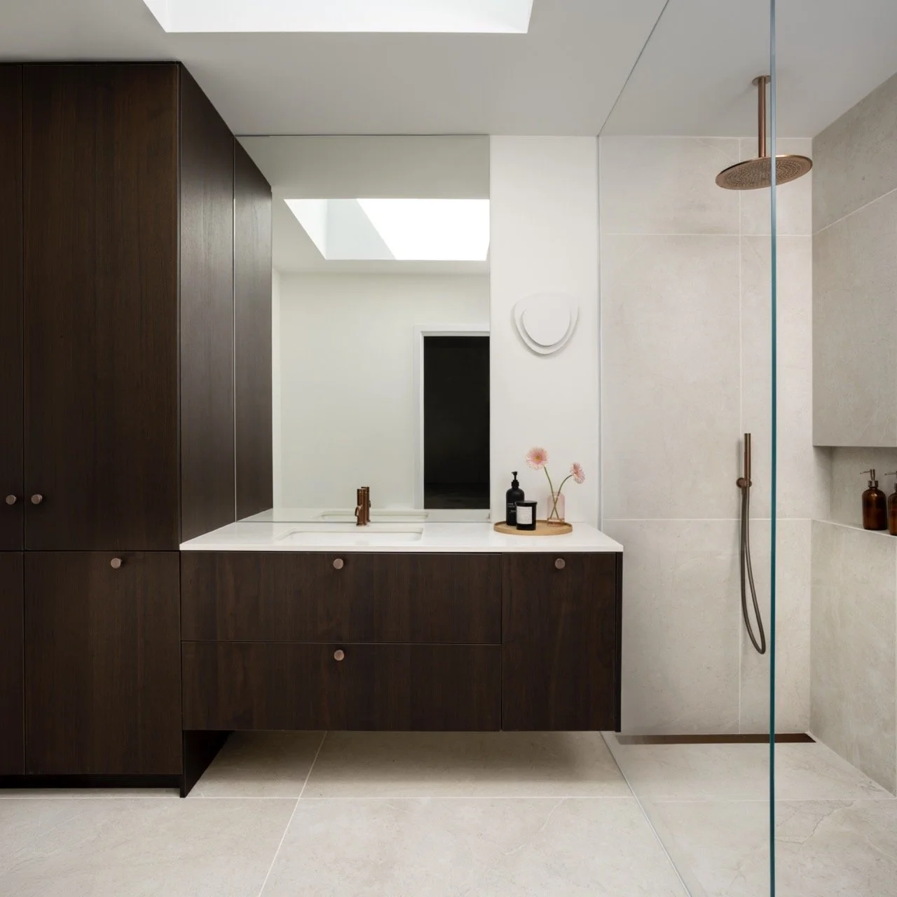 Modern bathroom with white and dark wood finishes, a large mirror, a shower area with a glass divider, and a minimalist decor with pink flowers and toiletries.