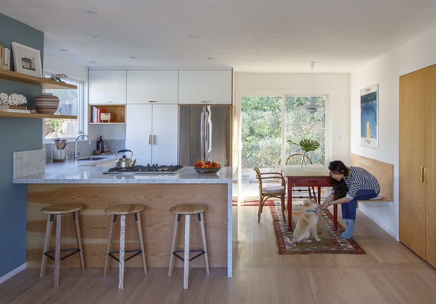 A woman sitting on a bench at a wooden dining table with a yellow Labrador puppy. The room has a bright kitchen with white cabinets, a marble countertop, and a large stainless steel refrigerator, with large glass doors opening to a garden.