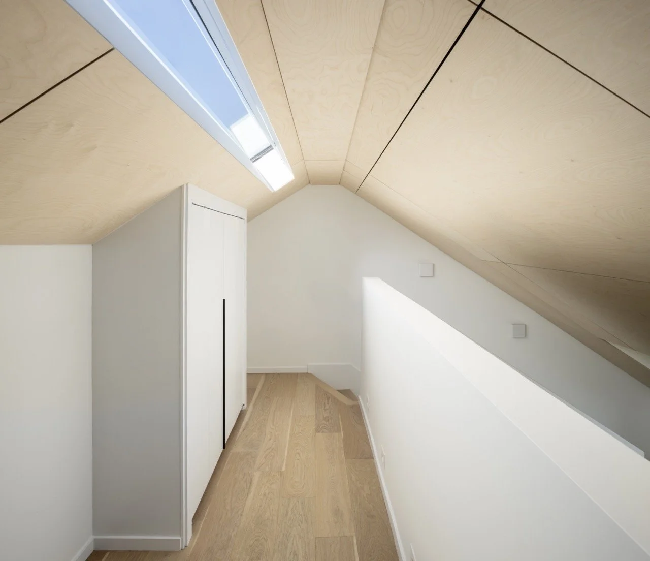 Small attic room with sloped wood-paneled ceiling, white walls, light wood flooring, a white cabinet, fluorescent light fixture, and minimal decor.