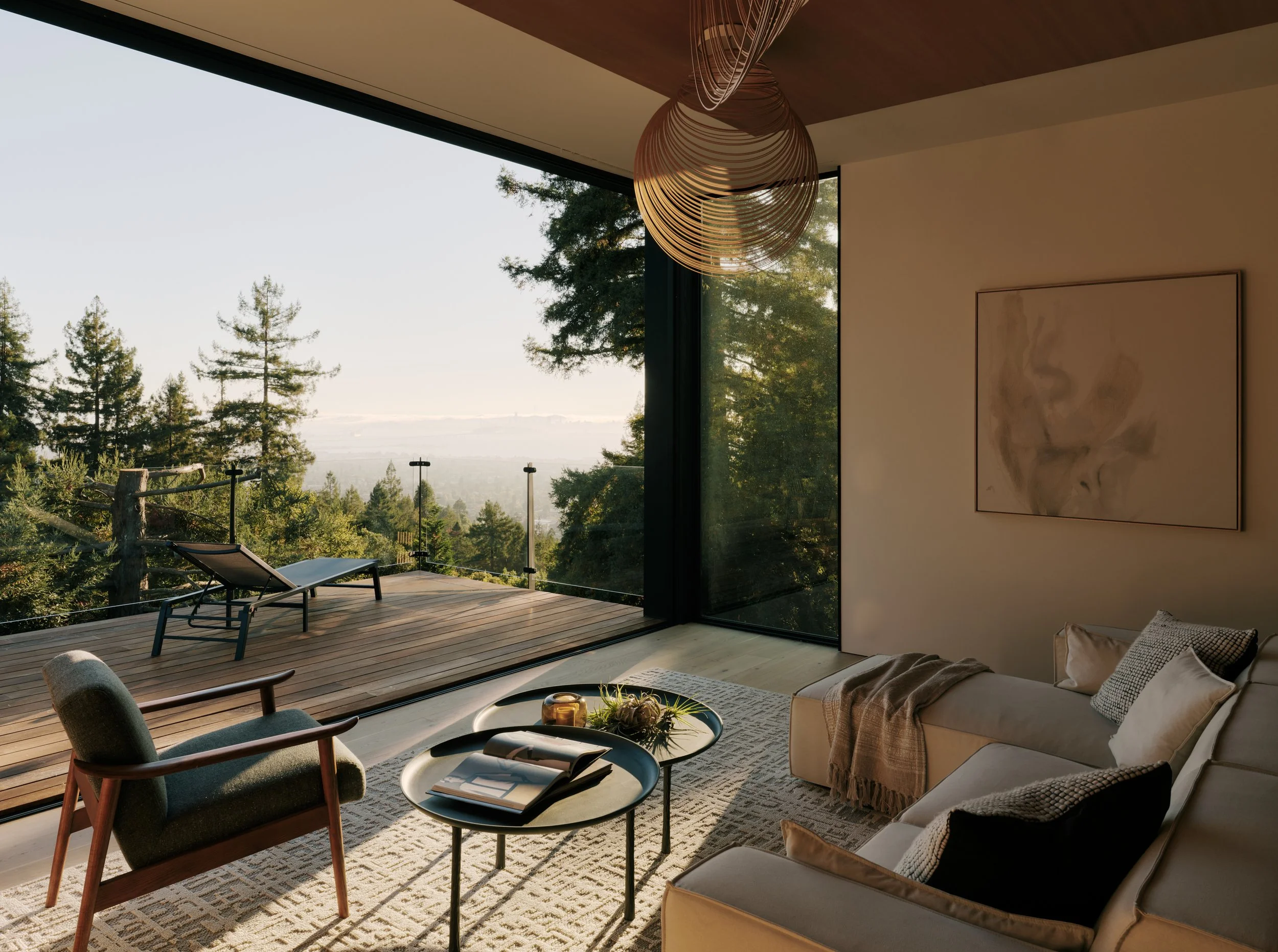 A modern living room with large floor-to-ceiling windows revealing a scenic view of trees and a landscape. The room features a beige sofa with throw pillows, a round coffee table with books and decor, and an outdoor deck with lounge chairs and a glas
