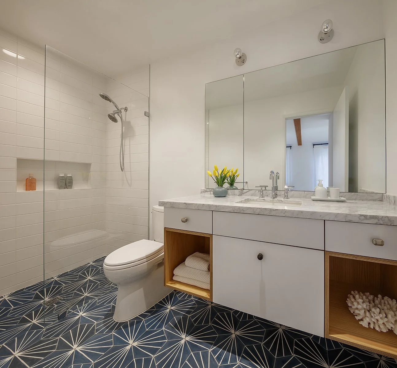 Modern bathroom with a walk-in shower, white tiles, a toilet, a marble countertop with a sink, a large mirror, yellow tulips, folded towels, soap, and decorative objects on open shelves, and patterned floor tiles.