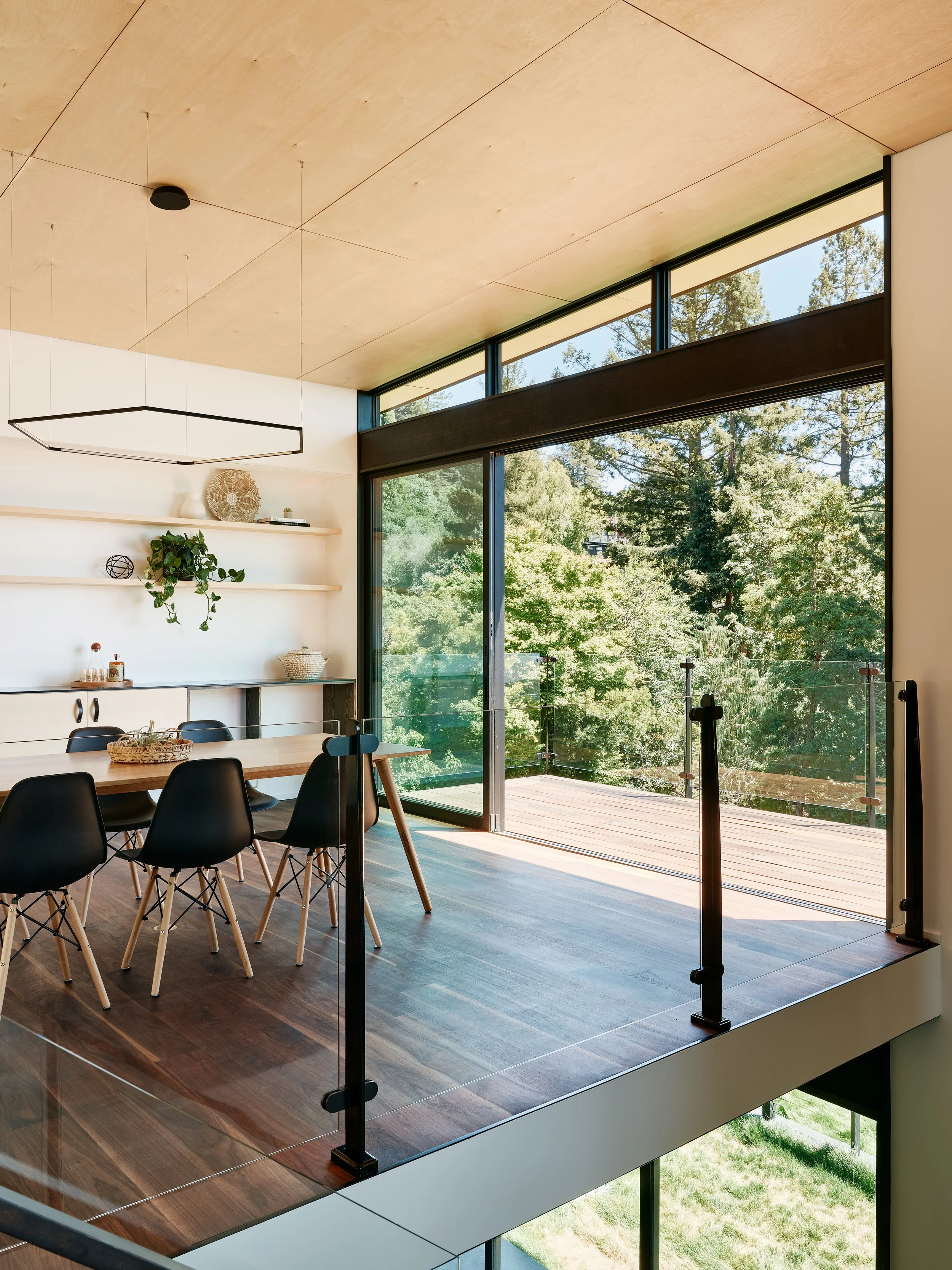 Modern, open-concept dining area with a rectangular wooden table, black chairs, and large glass balcony doors opening to a lush green outdoor view. The ceiling is wooden with a minimalist black chandelier, and a white wall with shelves holding decora