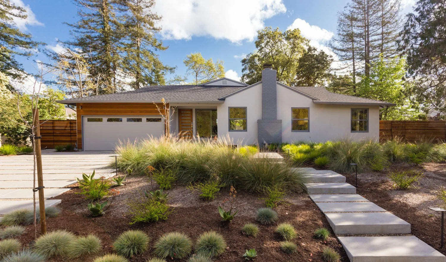 Modern single-story house with white exterior and gray chimney, surrounded by landscaped garden with various plants and trees, and a driveway leading to a garage.