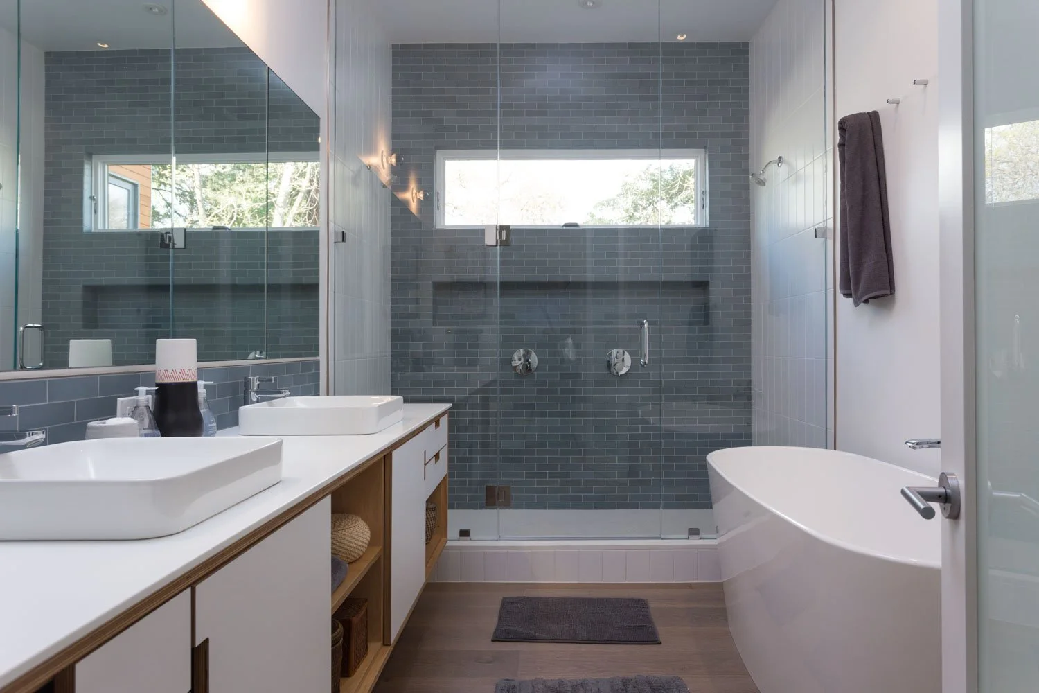 Modern bathroom with double sinks, a glass-enclosed shower with gray tile walls, a white bathtub, and a large mirror. Towel hangs on the wall and a small window lets in natural light.