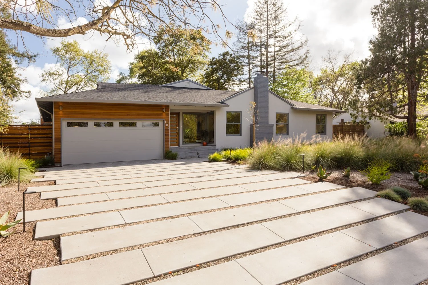 Front view of a modern house with a concrete driveway, landscaped yard, and large windows, surrounded by trees and a wooden fence.