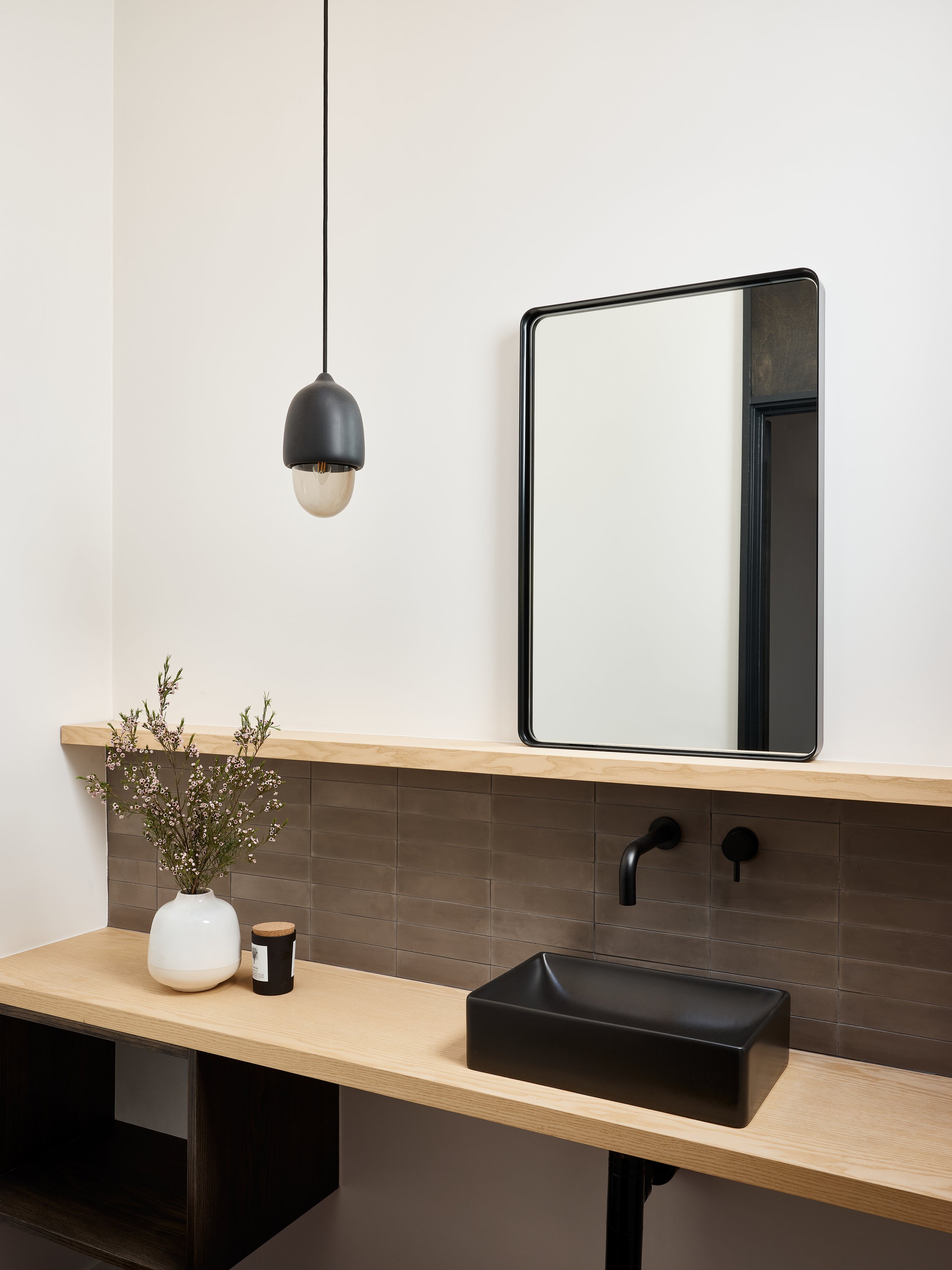 Modern bathroom with a black rectangular sink, a rectangular mirror, black faucet, wooden vanity with open shelving, a white vase with pink flowers on the countertop, and matching black mirror frame and pendant ceiling light.