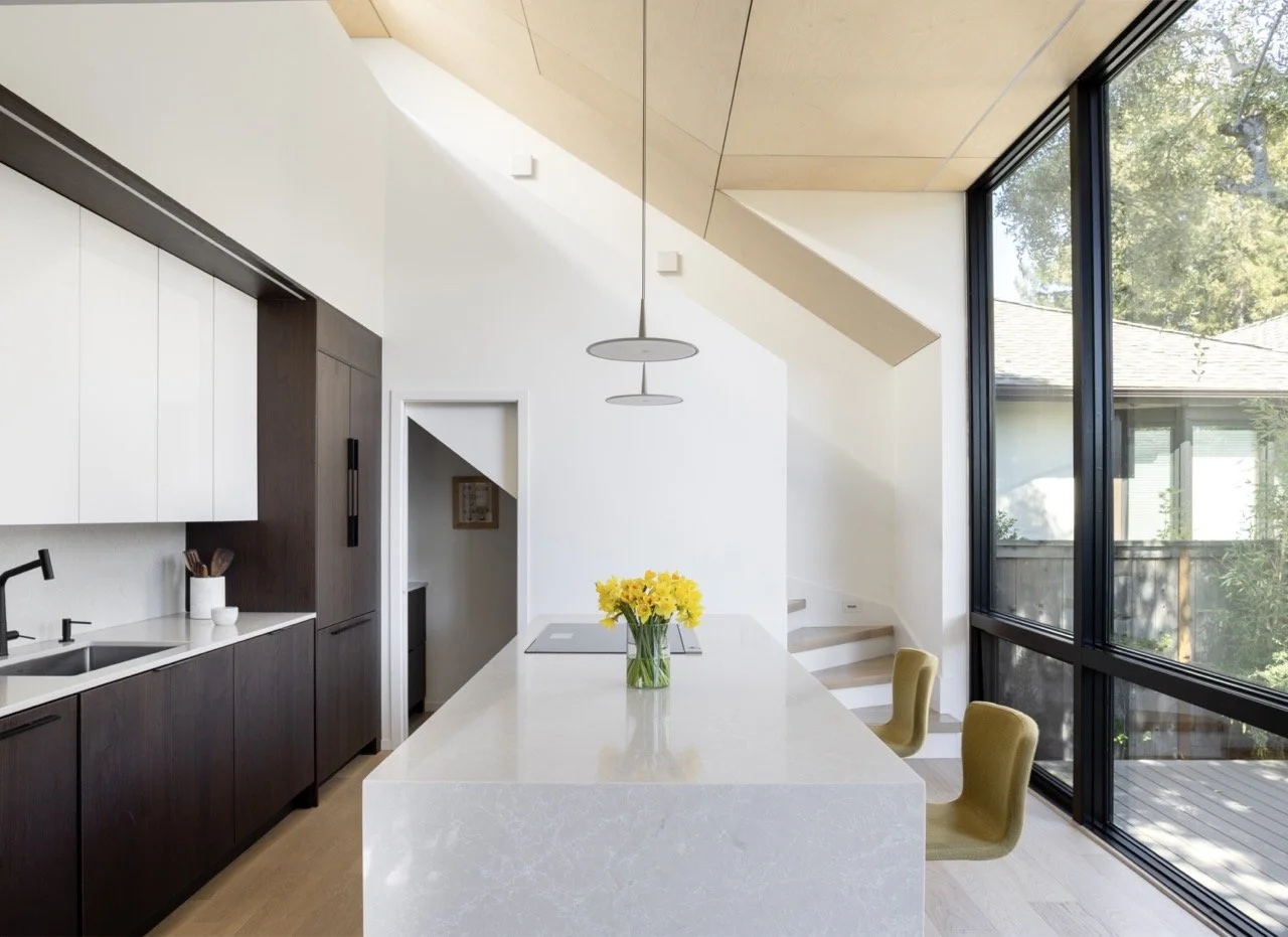 Modern kitchen with white and dark wood cabinets, a large island with a vase of yellow flowers, two olive-green chairs, large windows letting in natural light, and a view of a deck outside.