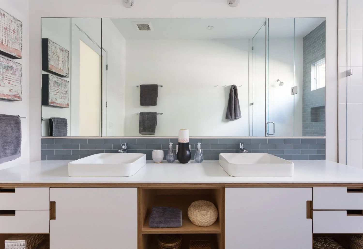 Modern bathroom with double white vessel sinks on a white countertop, mirror behind, gray subway tile backsplash, towels on the wall, and a glass shower with gray tile walls and a small window.