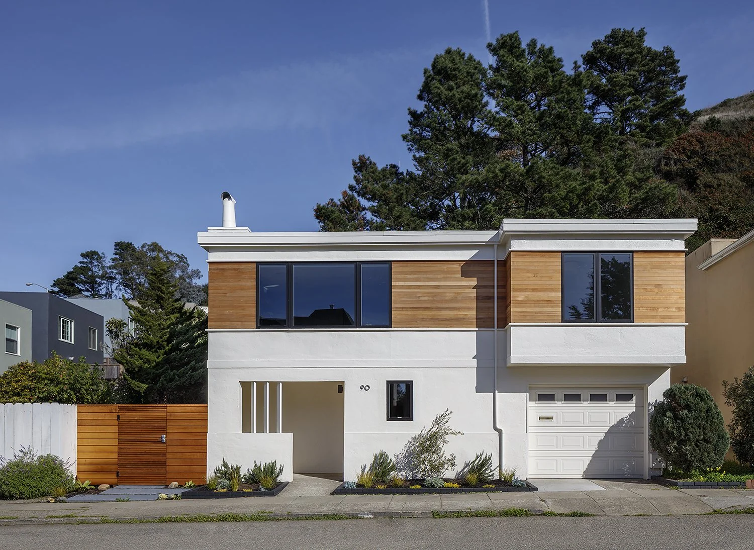 Modern two-story house with white and wood exterior, large upper windows, a garage, and landscaped front yard with plants and shrubs.