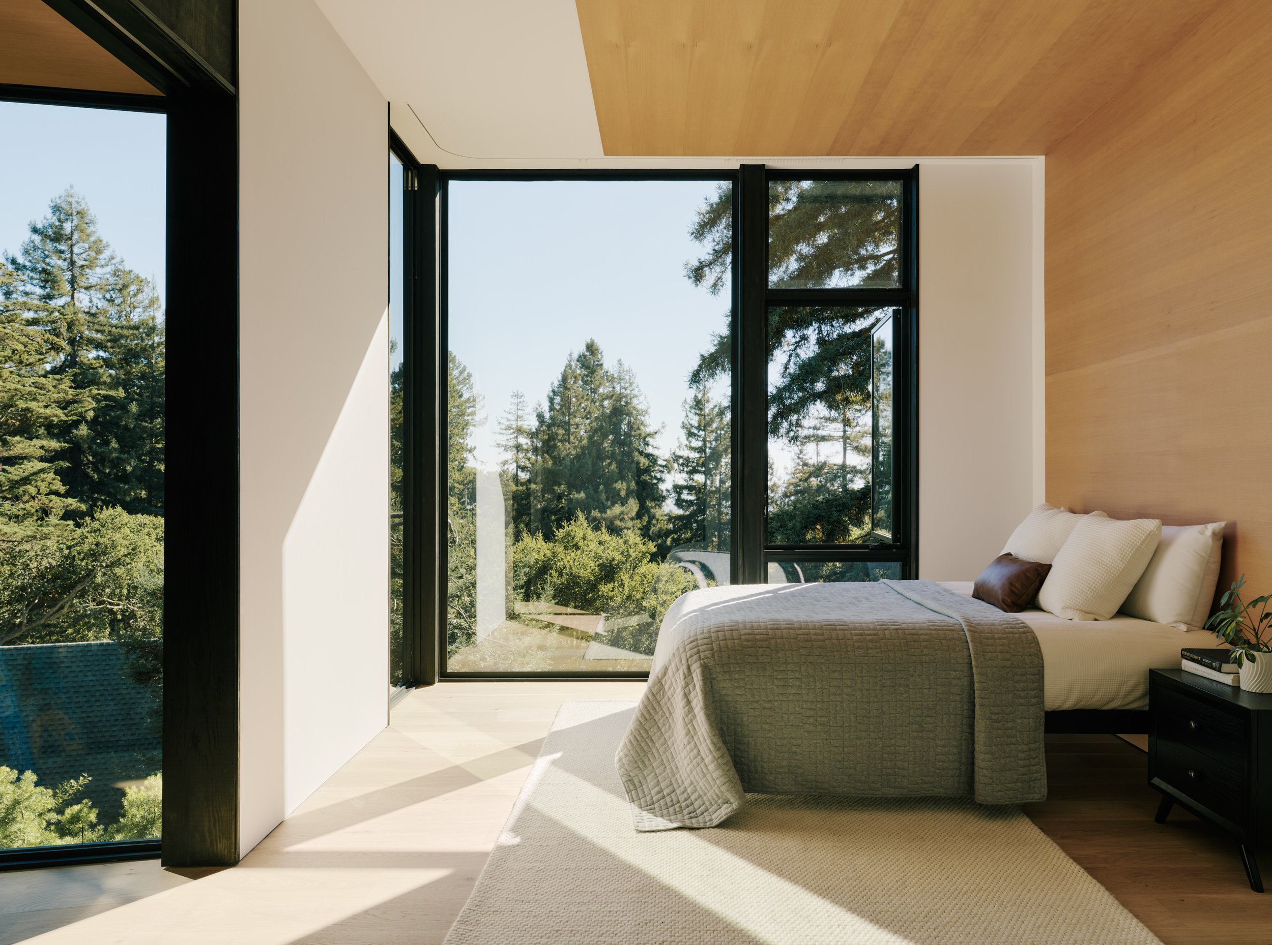 Modern bedroom with large floor-to-ceiling windows showing trees outside, white bedding, a dark nightstand with books and a small plant, and a wooden accent wall.