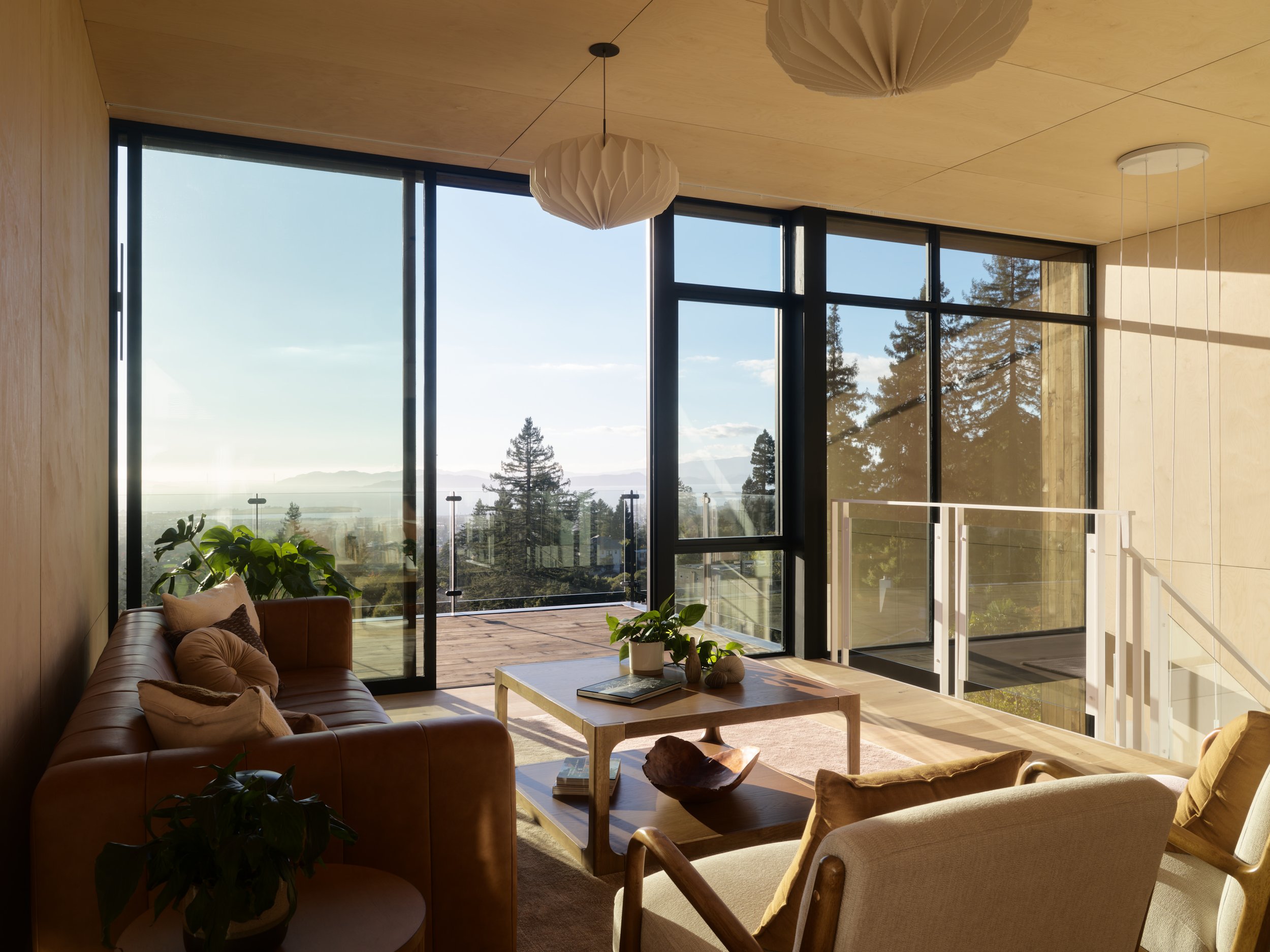 Living room with brown leather couch, beige armchair, wooden coffee table with plants and books, large floor-to-ceiling windows with a view of trees and mountains, wood-paneled ceiling, hanging light fixtures with paper shades.