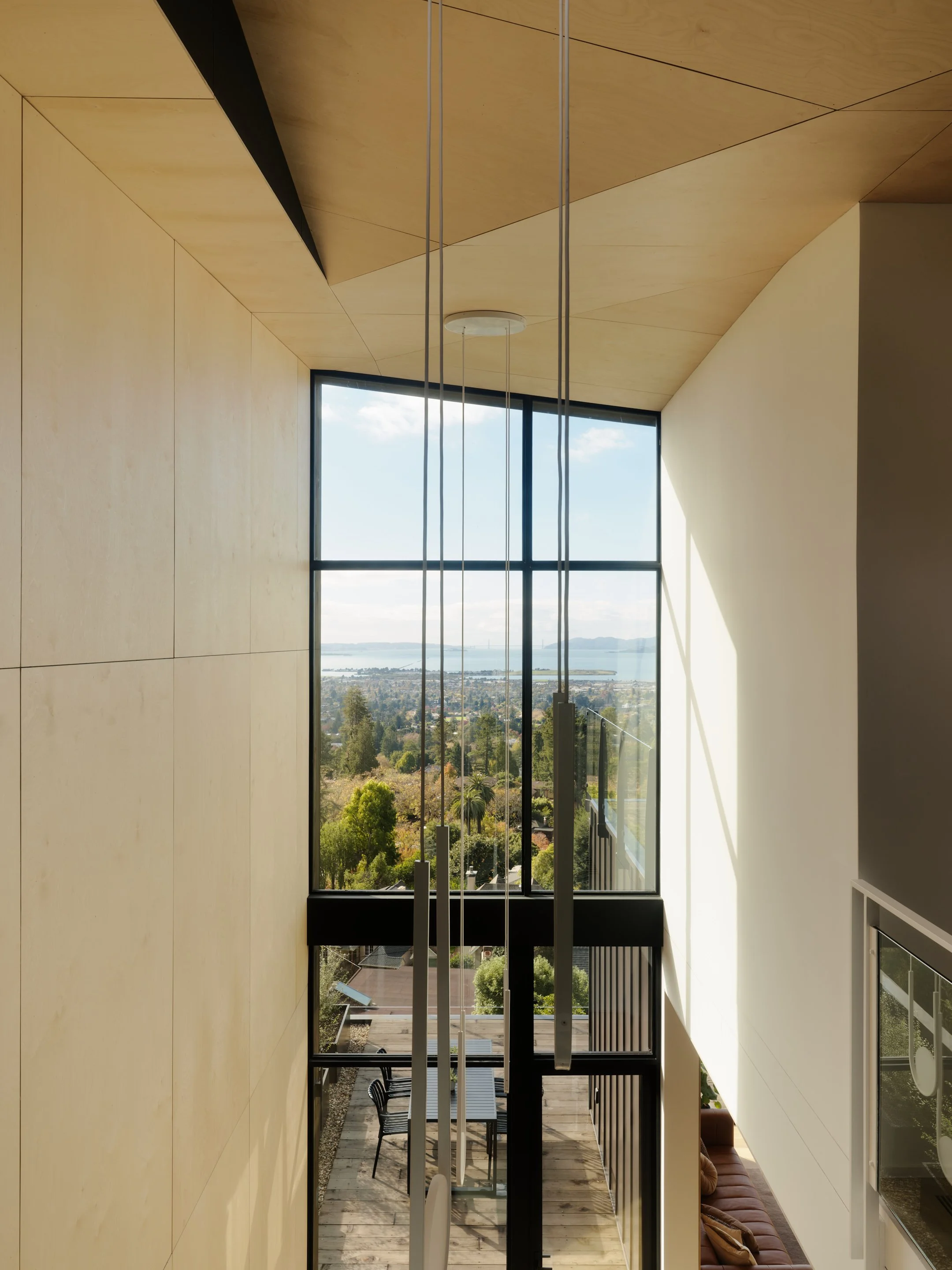 Modern interior with large floor-to-ceiling window overlooking a landscape with trees, chairs, and a table outside, high ceiling with hanging lights, and beige walls.