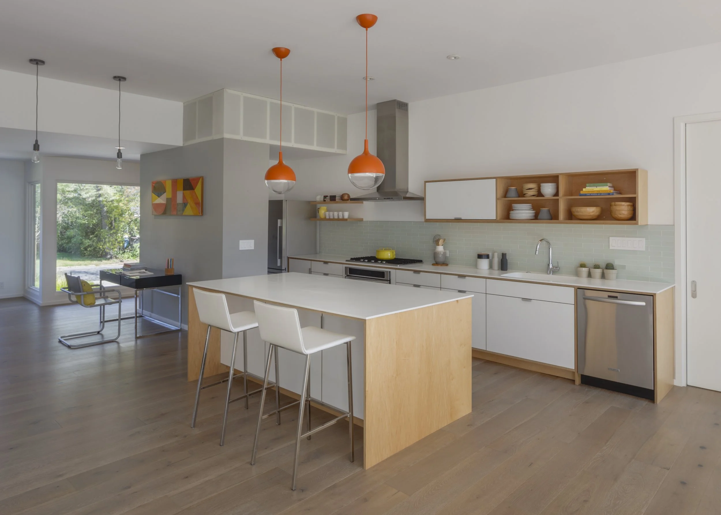 Modern kitchen with white cabinets, a wooden open shelf with dishes, a kitchen island with seating, two orange pendant lights, and large windows showing greenery outside.