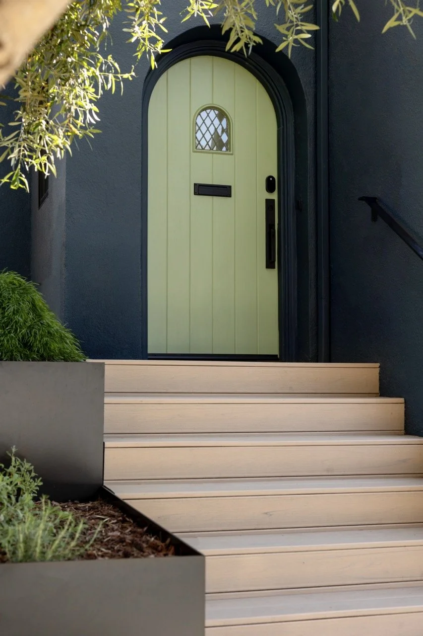 Front door with pale green color and small arched window, black mailbox, and black door hardware, accessed by wooden steps and surrounded by greenery.