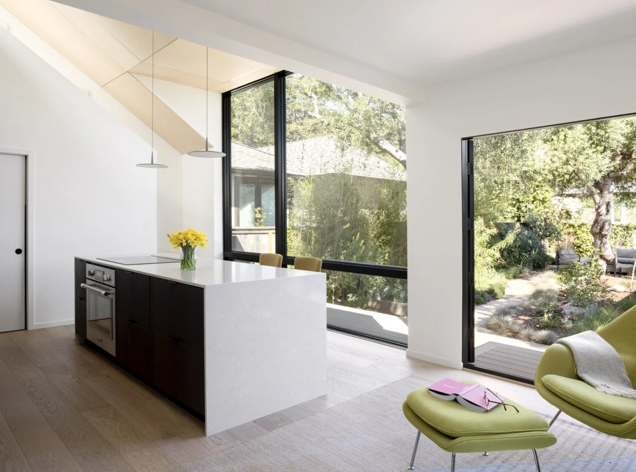 Modern kitchen with white walls, a kitchen island with a white countertop and dark cabinets, a vase of yellow flowers, pendant lights, large windows, and a sliding glass door leading to a garden with outdoor seating.